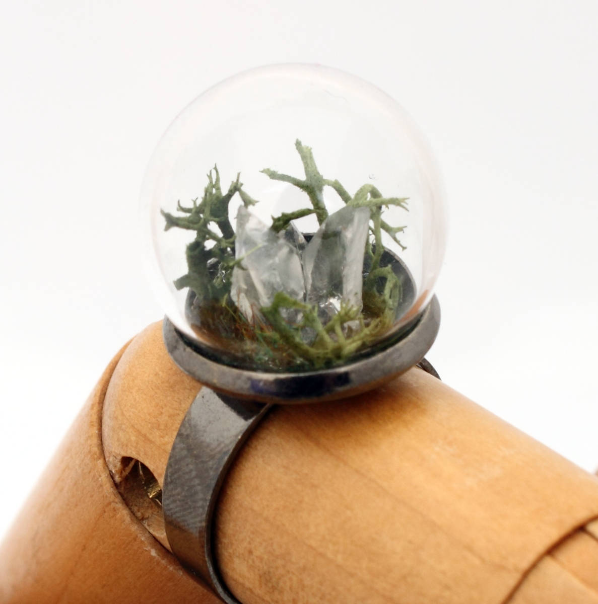 A wooden jointed hand displays this glass globe ring on a gunmetal ring base containing a crystal of lemurian seed quartz. The quartz is accompanied by reindeer moss and greenery inside the dome and displayed against a white background