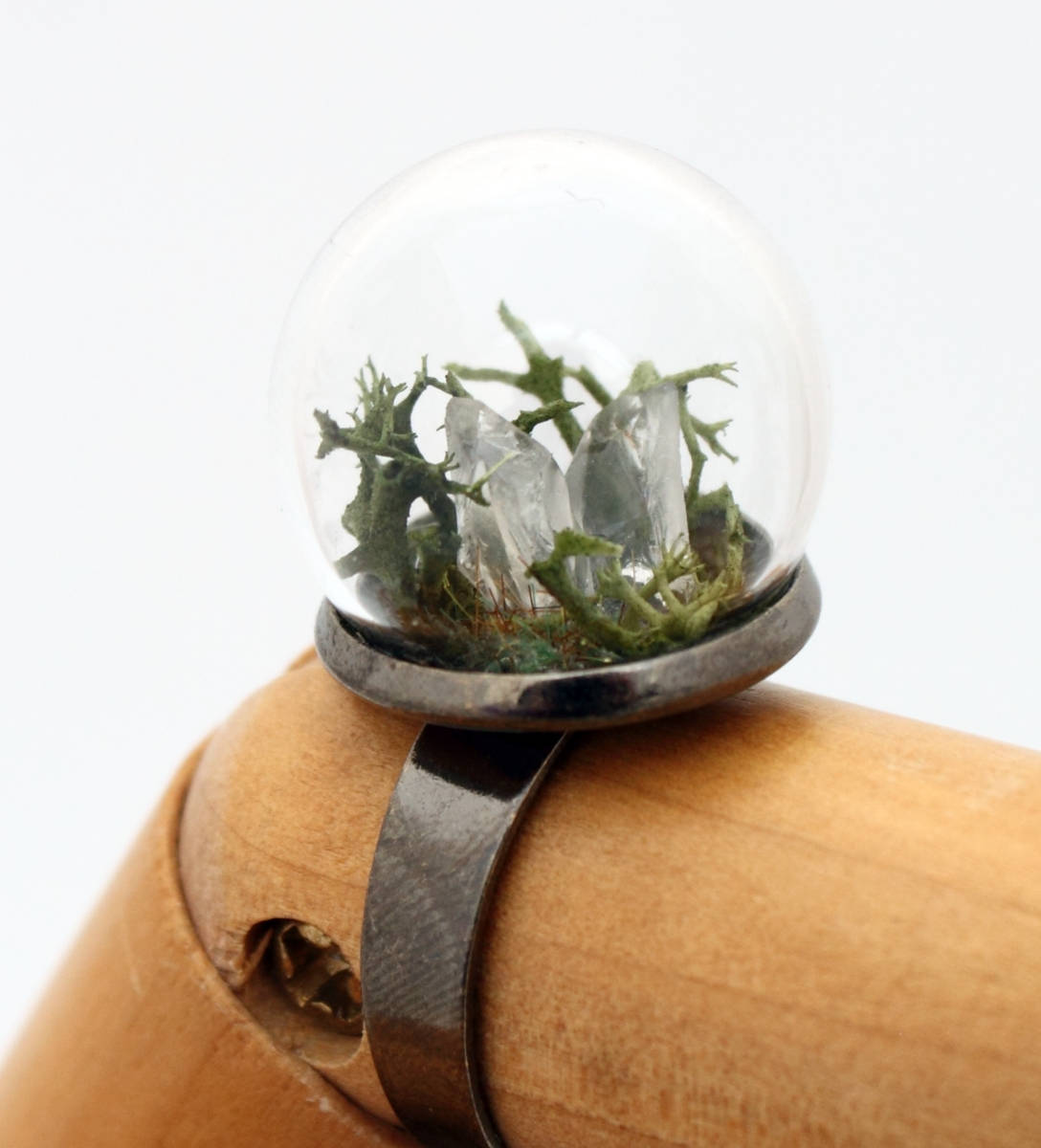 A wooden jointed hand displays this glass globe ring on a gunmetal ring base containing a crystal of lemurian seed quartz. The quartz is accompanied by reindeer moss and greenery inside the dome and displayed against a white background