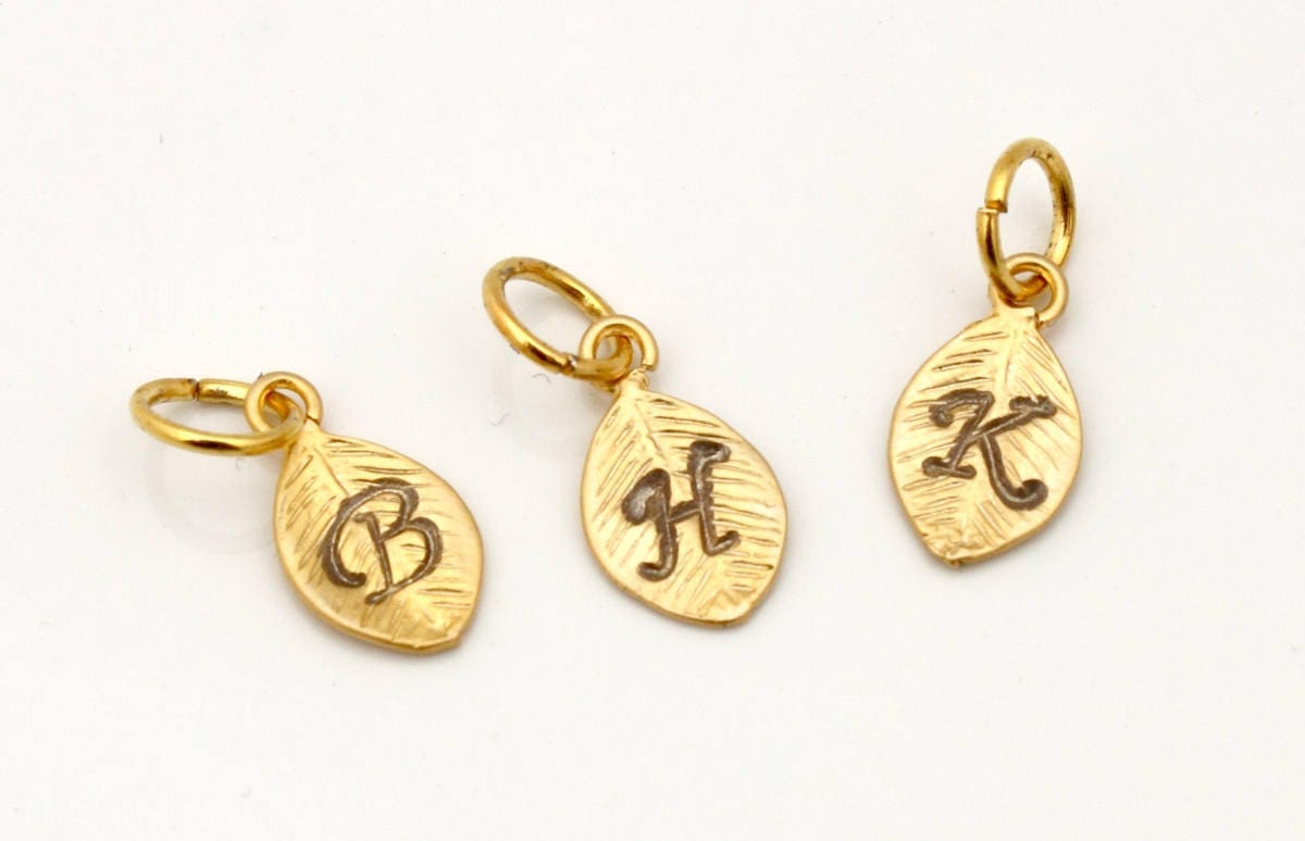 3 metal leaf-shaped charms with a small loop and jump ring on a white background. The charms are gold coloured with a stamped calligraphic initials B, H and K