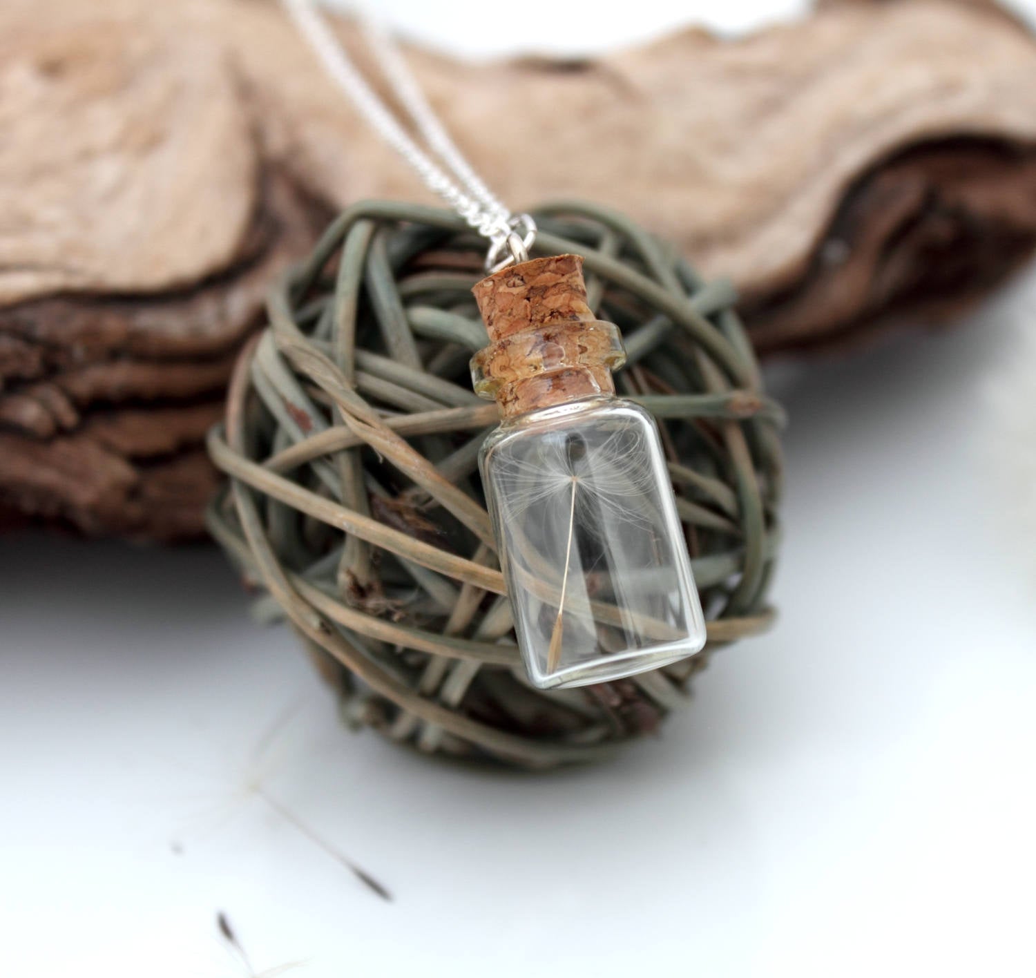 A glass vial necklace contains a single dandelion seed. The dandelion vial hangs on a fine sterling silver chain and is displayed on a grey wicker ball