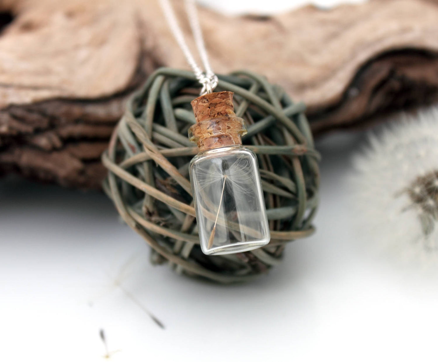 A glass vial necklace contains a single dandelion seed. The dandelion vial hangs on a fine sterling silver chain and is displayed on a grey wicker ball