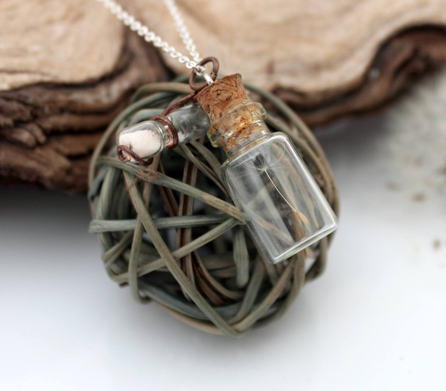 Miniature jar shaped glass vial necklace containing a dandelion seed with a copper wrapped miniature glass sand timer attachdd to the cork lid. Displayed on a silver coloured chain attached to the cork lid