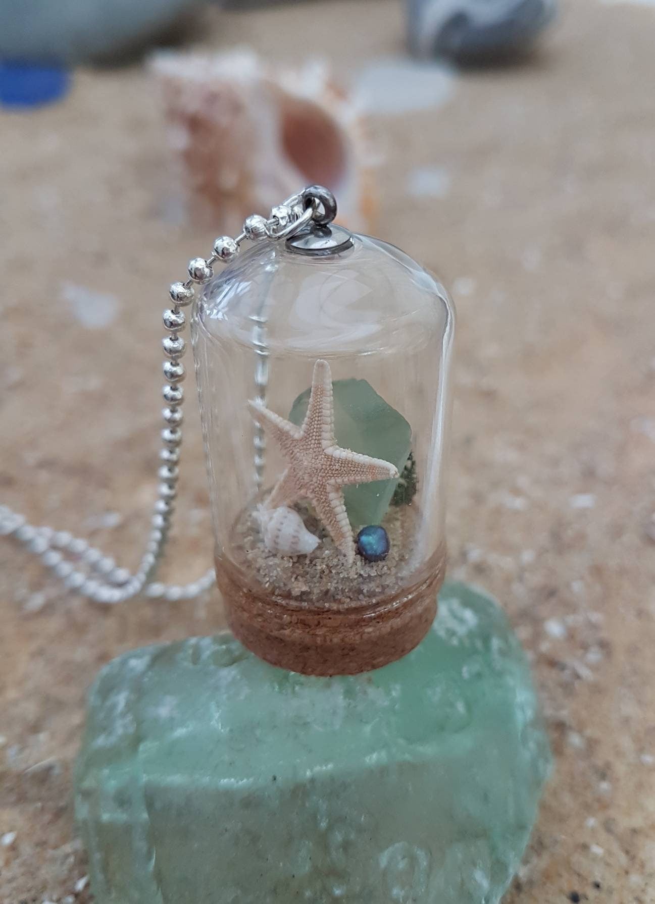 Small glass bell jar with cork base and necklace attachment containing a starfish, shell, pearl, light green seaglass and greenery. It sits on a piece of aquamarine seaglass and sand