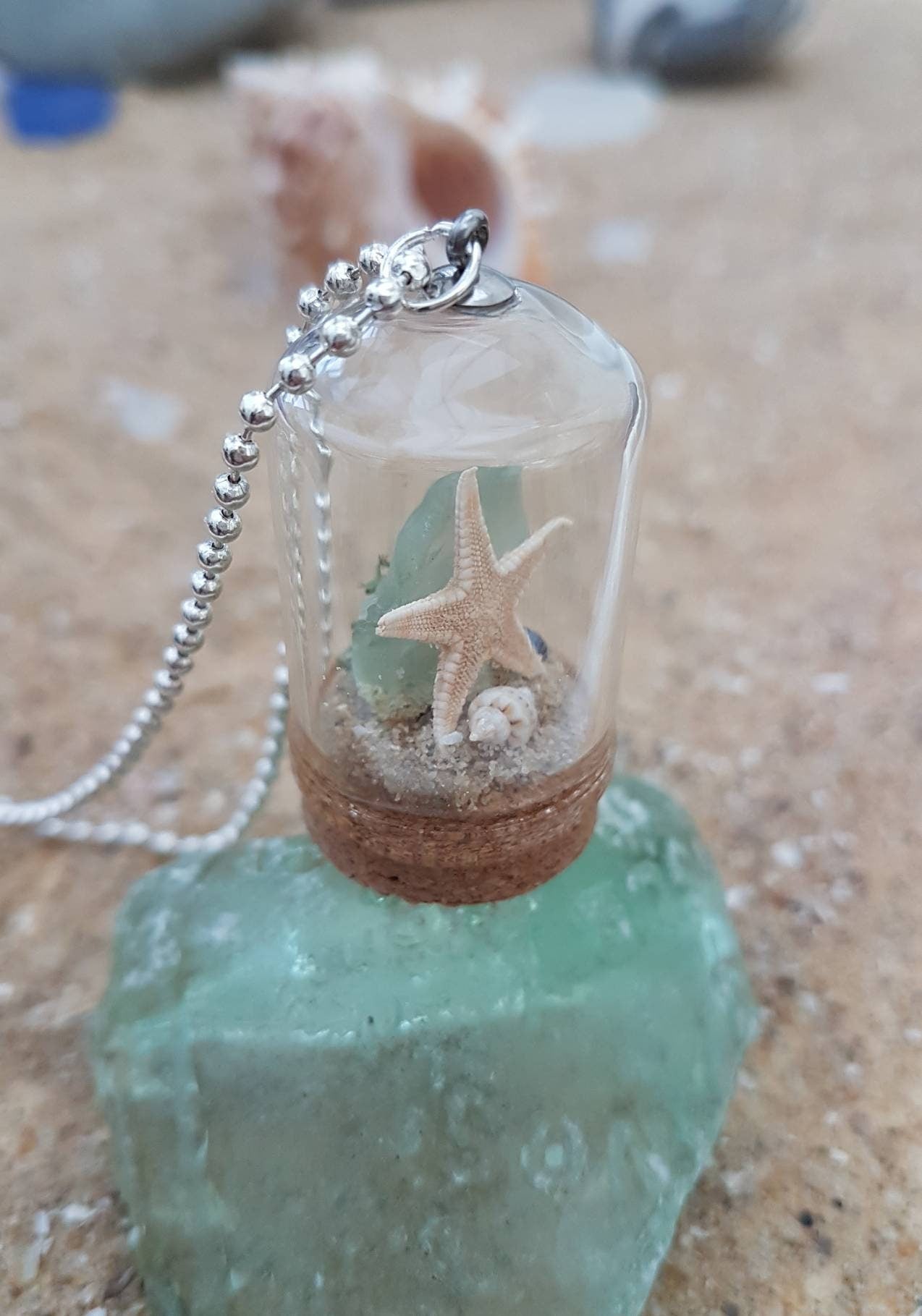 Small glass bell jar with cork base and necklace attachment containing a starfish, shell, pearl, light green seaglass and greenery. It sits on a piece of aquamarine seaglass and sand