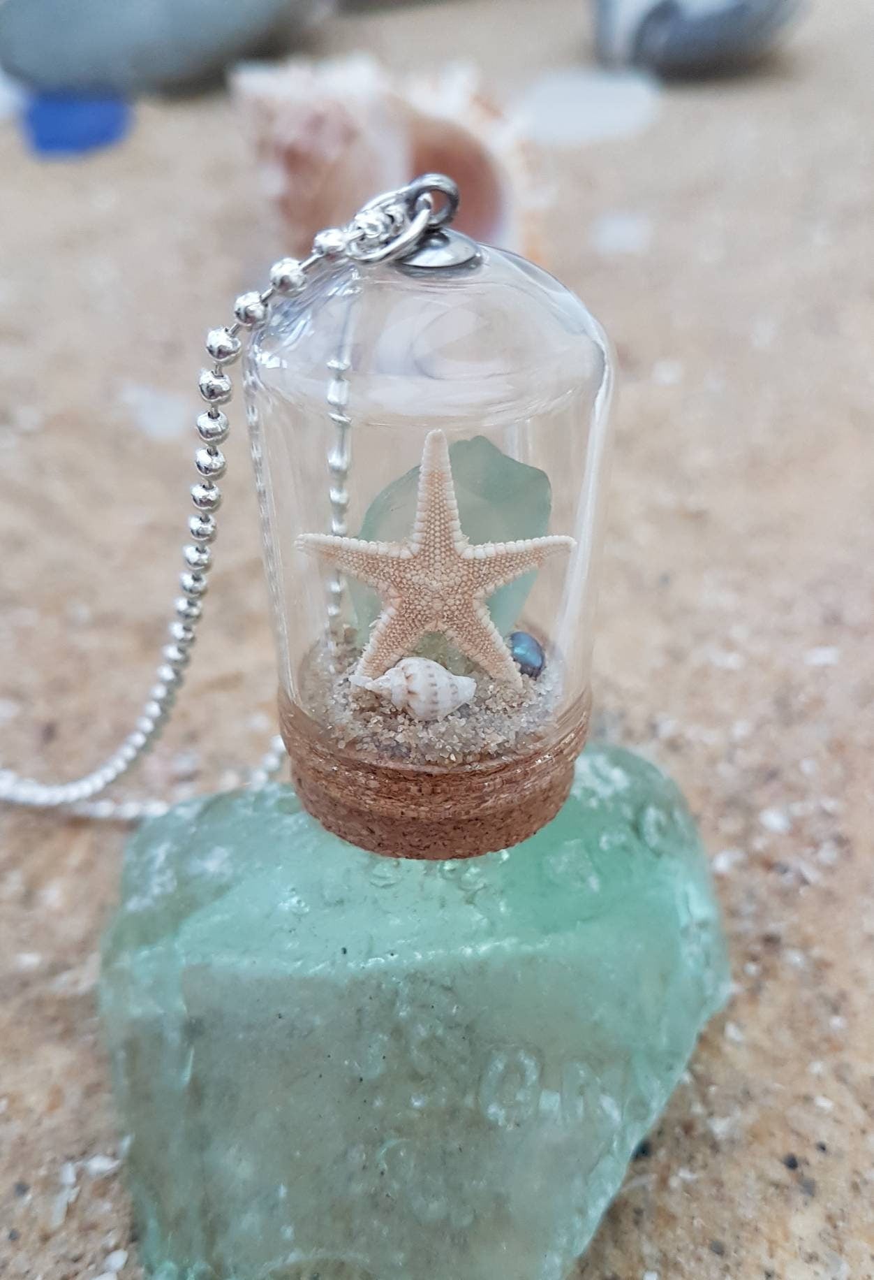 Small glass bell jar with cork base and necklace attachment containing a starfish, shell, pearl, light green seaglass and greenery. It sits on a piece of aquamarine seaglass and sand