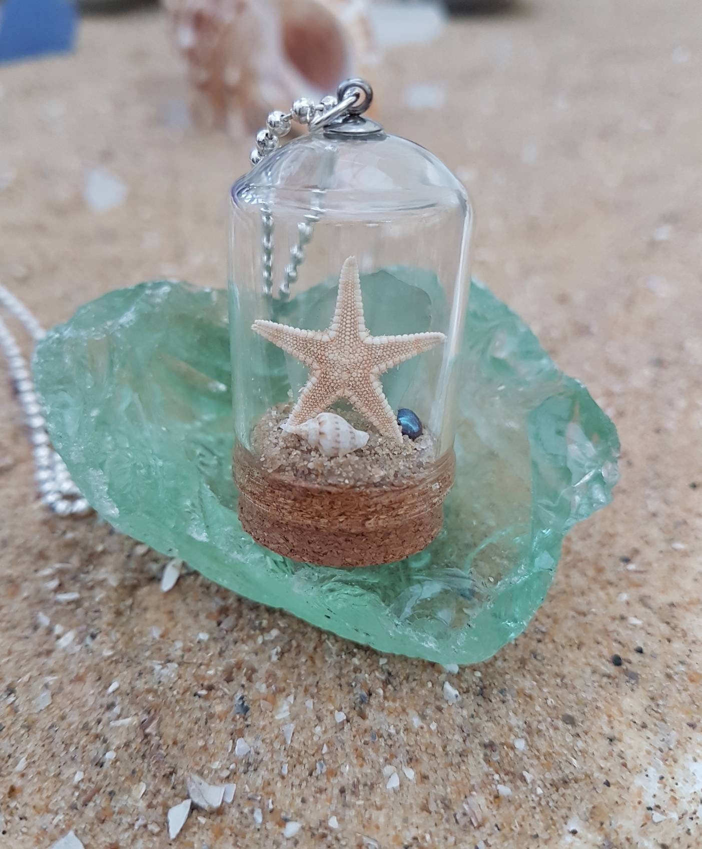 Small glass bell jar with cork base and necklace attachment containing a starfish, shell, pearl, light green seaglass and greenery. It sits on a piece of aquamarine seaglass and sand