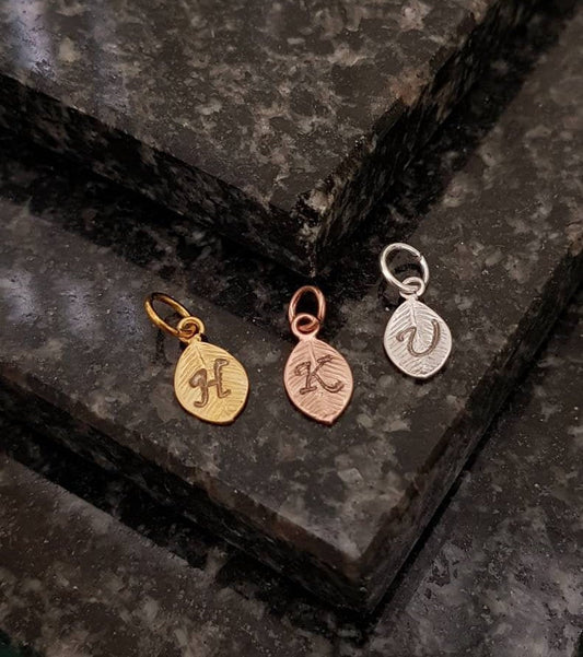 3 metal leaf-shaped charms with a small loop and jump ring. The first is gold coloured with a stamped calligraphic H initial. The second is rose gold coloured with a K initial. The third is silver coloured with a V initial. Black granite background