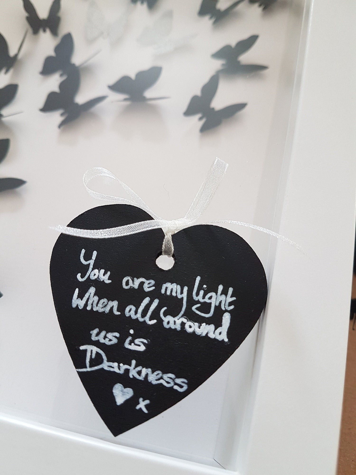 A small black heart is shown with silver writing that reads You are my light when all around us is darkness x. The heart is shown resting on the plastic facade of the white box frame. Black 3D butterflies is seen in the background