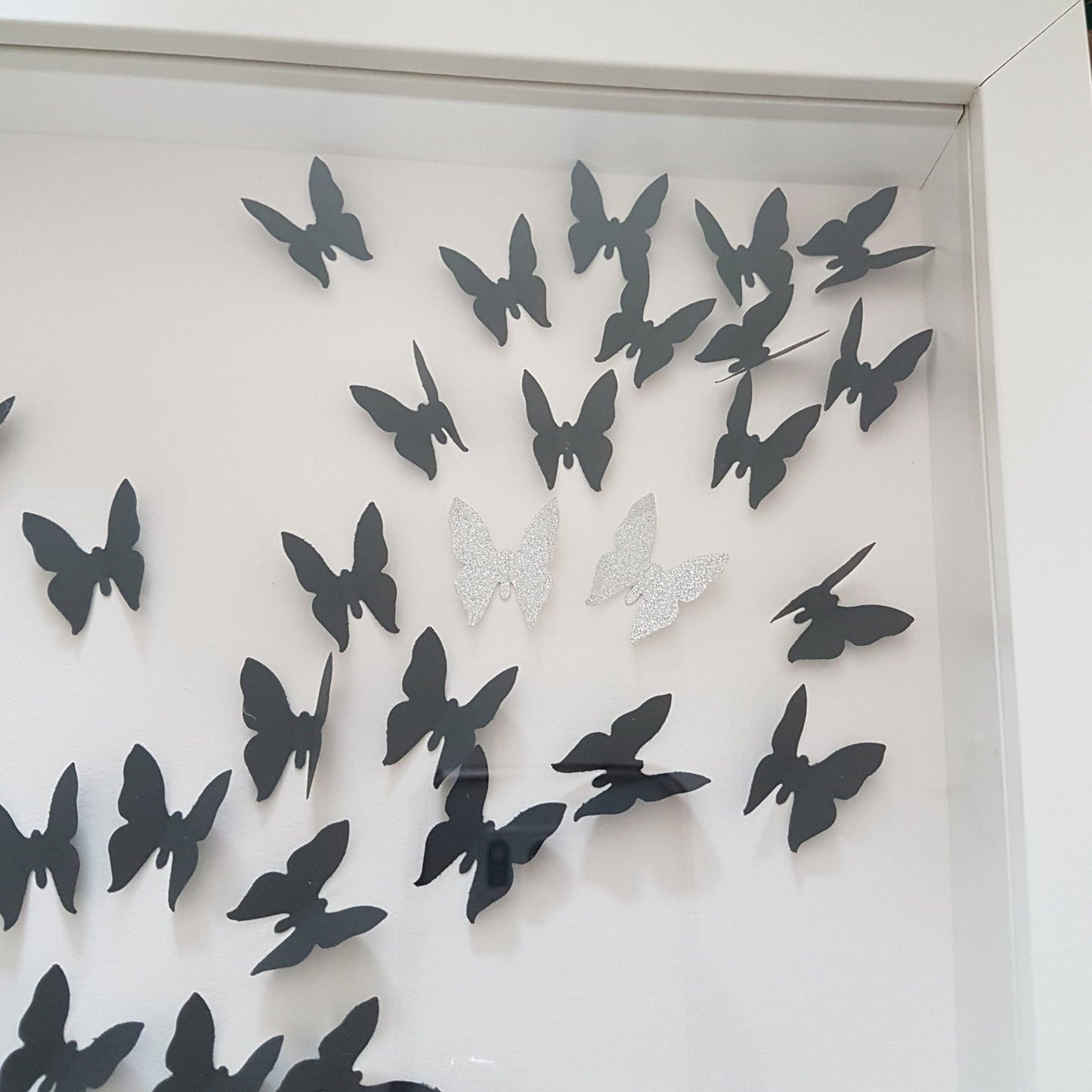 Square white box frame showing a flurry of 3D black paper butterflies with two silver glitter butterflies, moving from the bottom left of the frame, through to the top right of the frame.  Shown against a white background