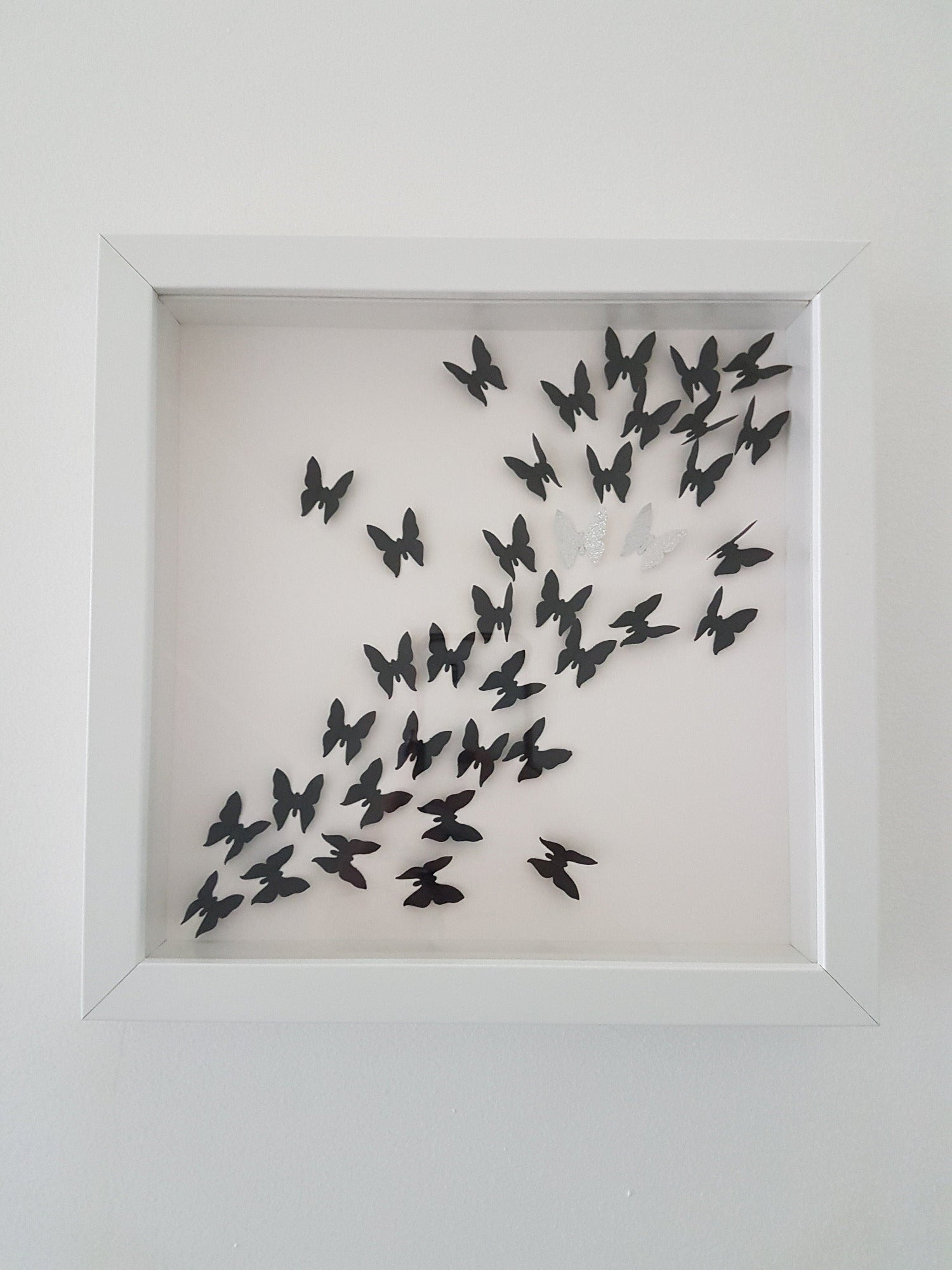 Square white box frame showing a flurry of 3D black paper butterflies with two silver glitter butterflies, moving from the bottom left of the frame, through to the top right of the frame.  Shown against a white background