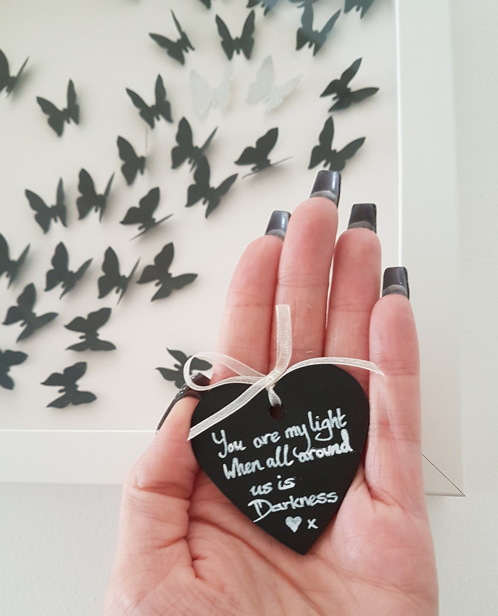 Open palm of a hand shown holding a small black heart with silver writing that reads You are my light when all around us is darkness x. The white box frame with black 3D butterflies is seen in the background