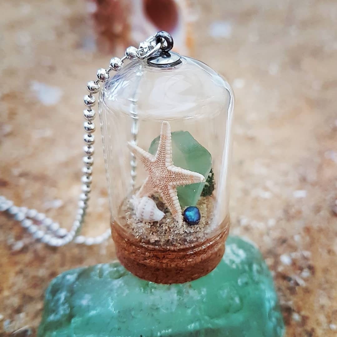 Small glass bell jar with cork base and necklace attachment containing a starfish, shell, pearl, light green seaglass and greenery. It sits on a piece of aquamarine seaglass and sand