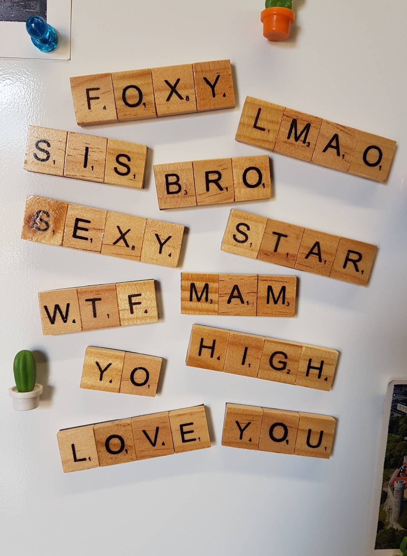 White fridge door displays 12 rectangular wooden scrabble tile magnets. The words shown include Foxy, Sis, bro, love, you, WTF, LMAO