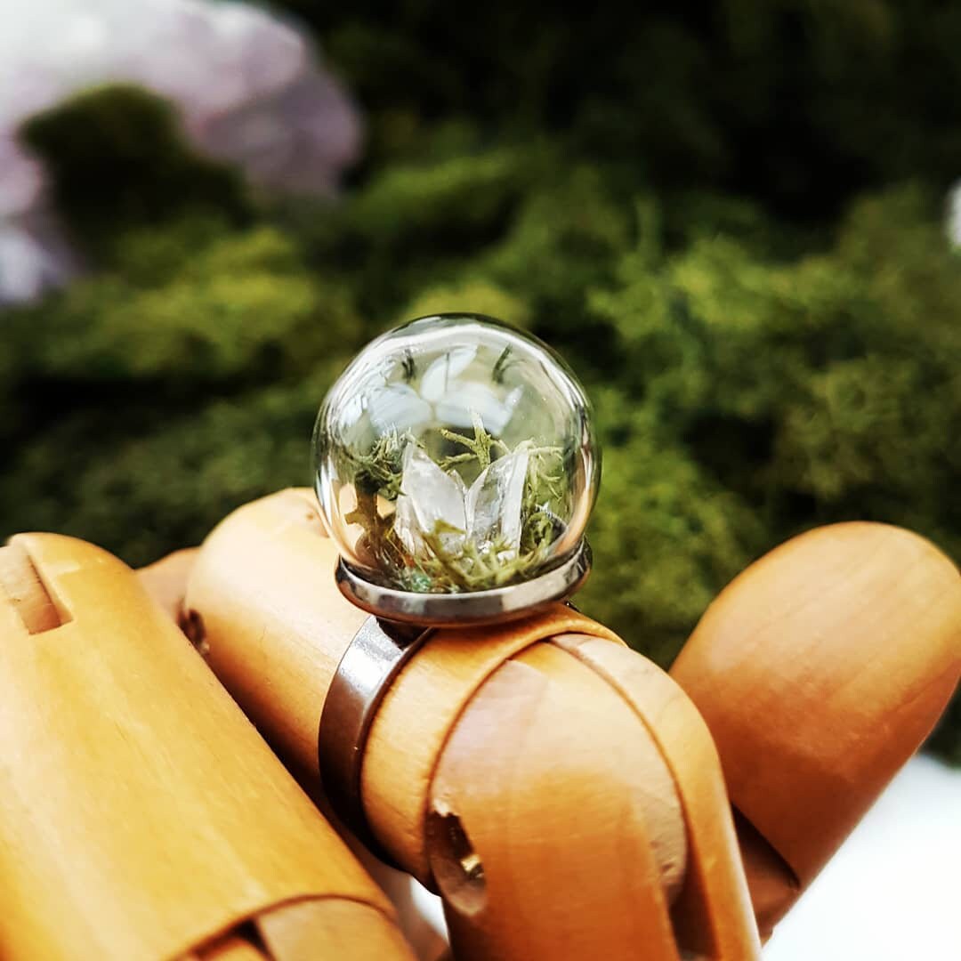 A wooden jointed hand displays this glass globe ring on a gunmetal ring base containing a crystal of lemurian seed quartz. The quartz is accompanied by reindeer moss and greenery inside the dome and displayed against a green background