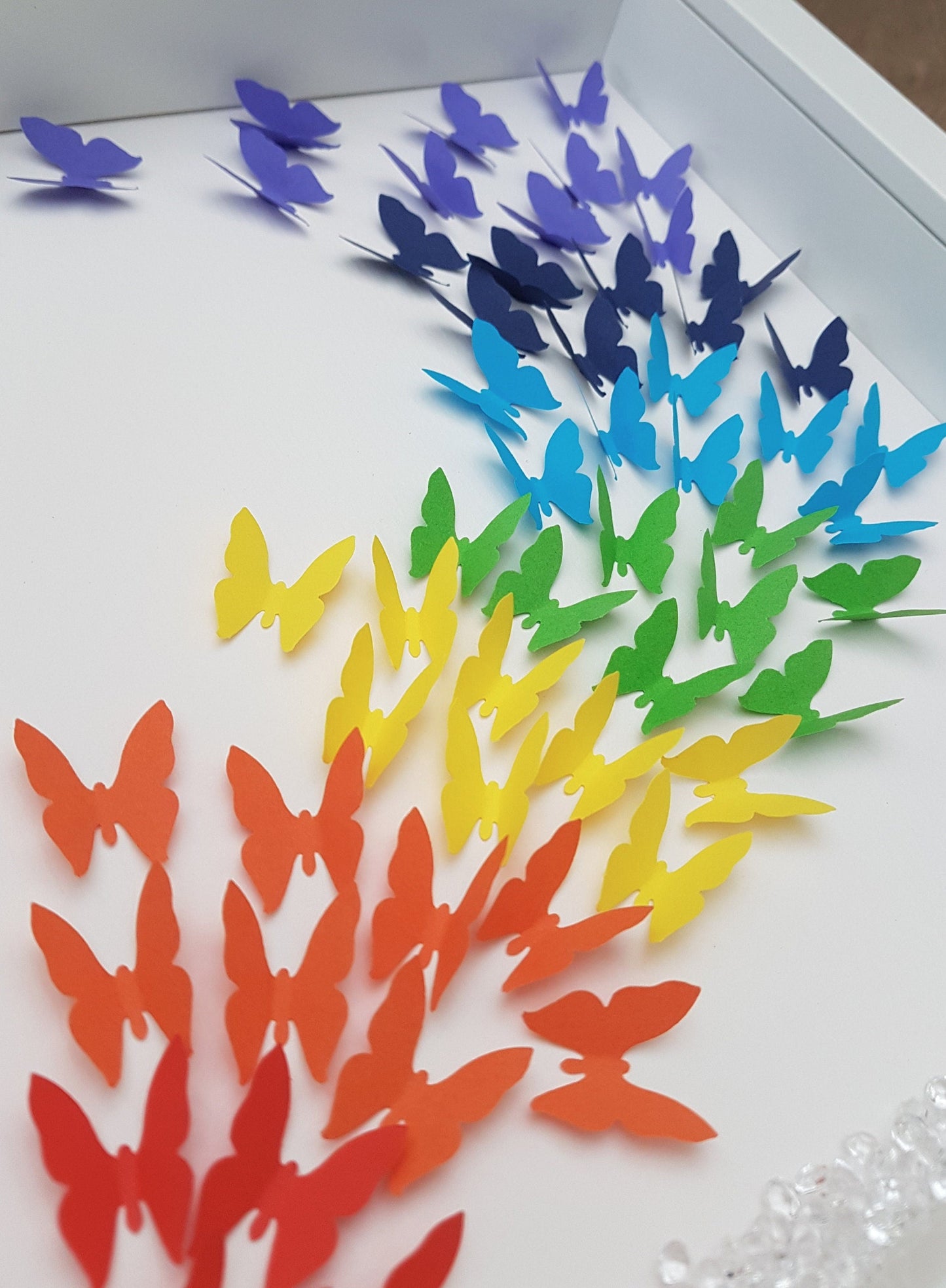 Square white box frame showing a flurry of paper butterflies in rainbow colours, moving from red butterflies in the bottom left of the frame, through rainbow colours to the top right of the frame. Detail shows red, orange and yellow butterflies