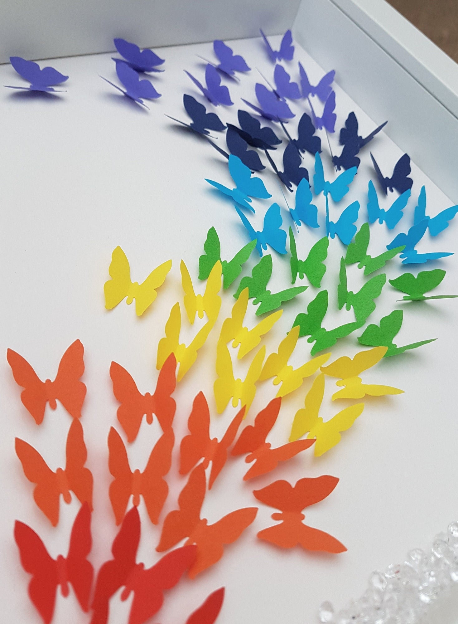 Square white box frame showing a flurry of paper butterflies in rainbow colours, moving from red butterflies in the bottom left of the frame, through rainbow colours to the top right of the frame. Detail shows red, orange and yellow butterflies