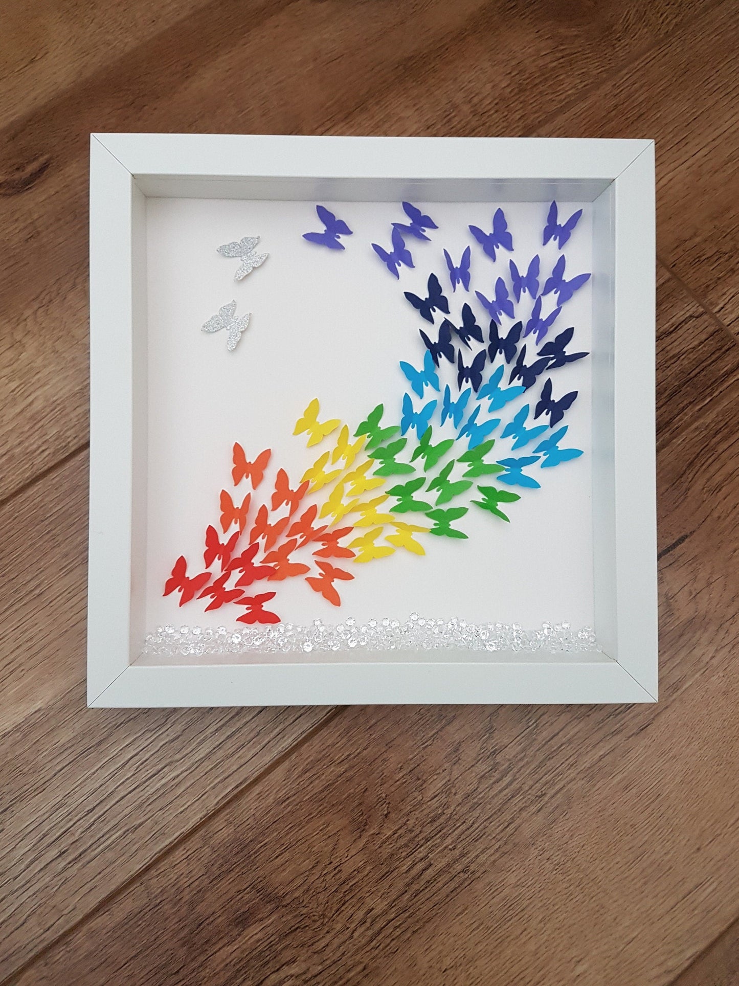 Square white box frame showing a flurry of paper butterflies in rainbow colours, moving from red butterflies in the bottom left of the frame, through rainbow colours to the top right of the frame.  Shown against a wooden background