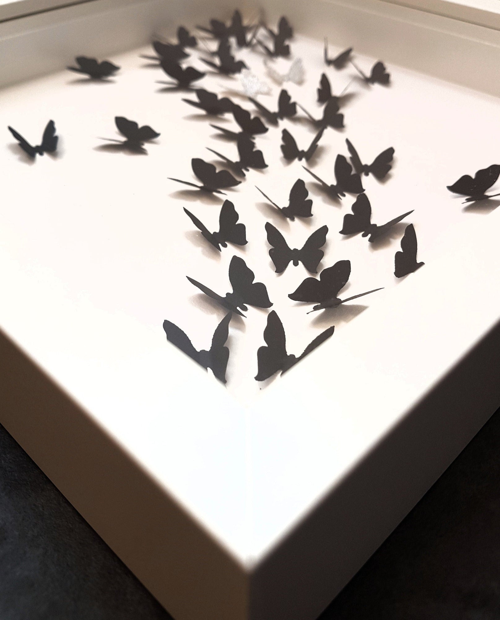 Square white box frame showing a flurry of black paper 3D butterflies with two silver glitter butterflies, moving from the bottom left of the frame, through to top right. Shown against a white background from an angle to show 3D nature of butterflies