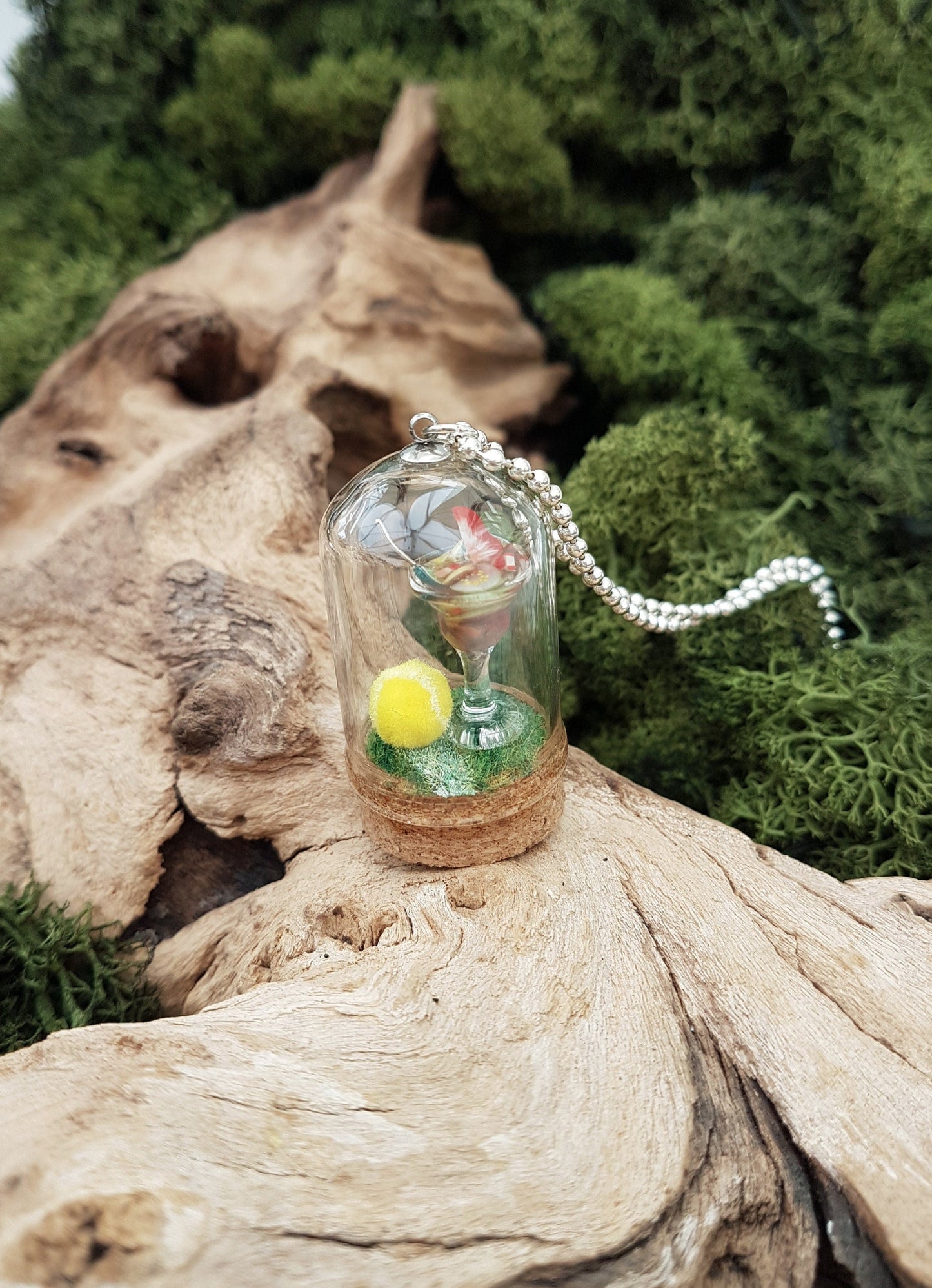 Miniature glass bell jar containing a miniature cocktail glass with Pimms and miniature fruit slices sat on a green flocked base with a fluorescent yellow tennis ball. A silver coloured chain is attached to the top of the bell jar