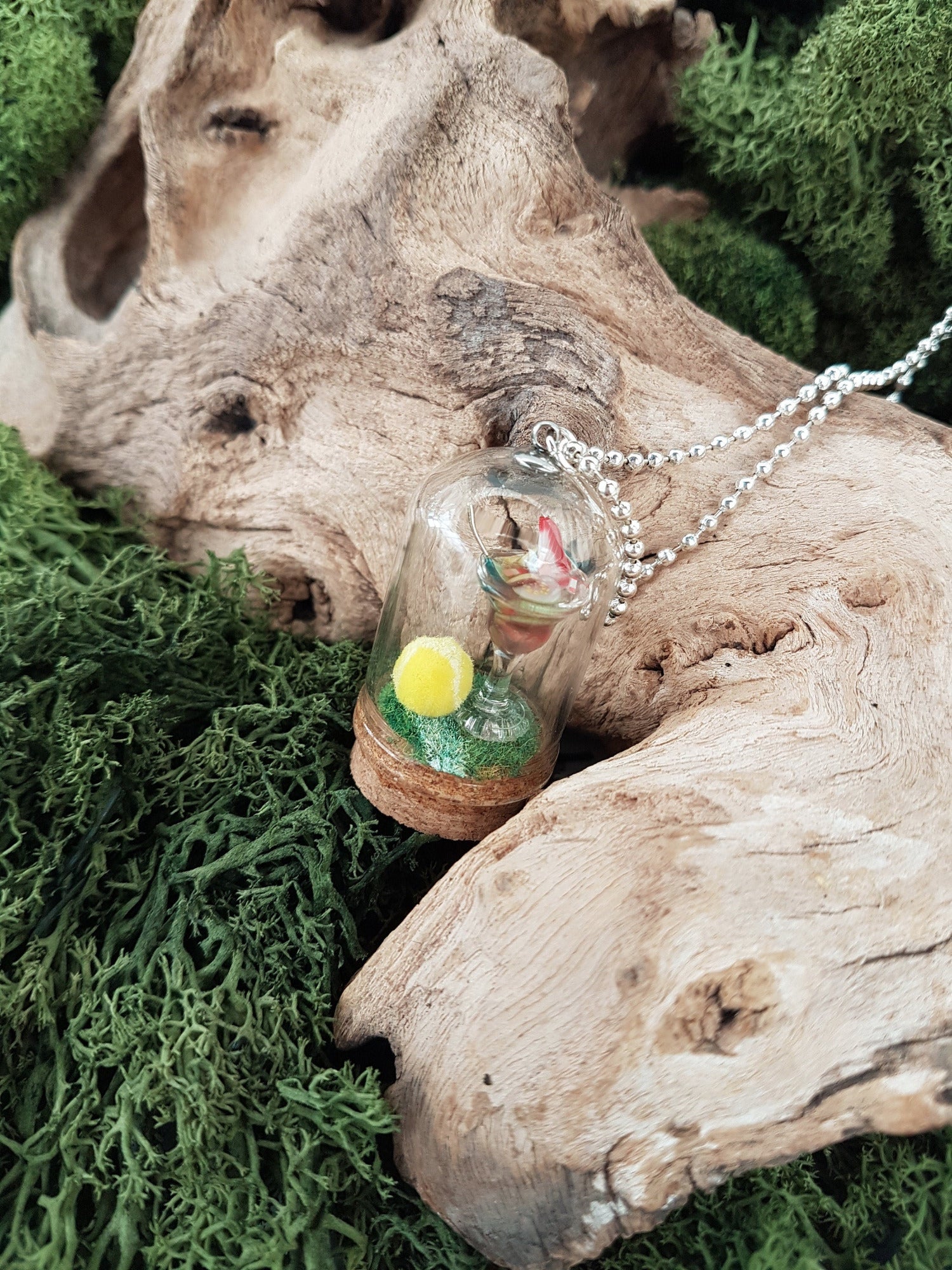 Miniature glass bell jar containing a miniature cocktail glass with Pimms and miniature fruit slices sat on a green flocked base with a fluorescent yellow tennis ball. A silver coloured chain is attached to the top of the bell jar