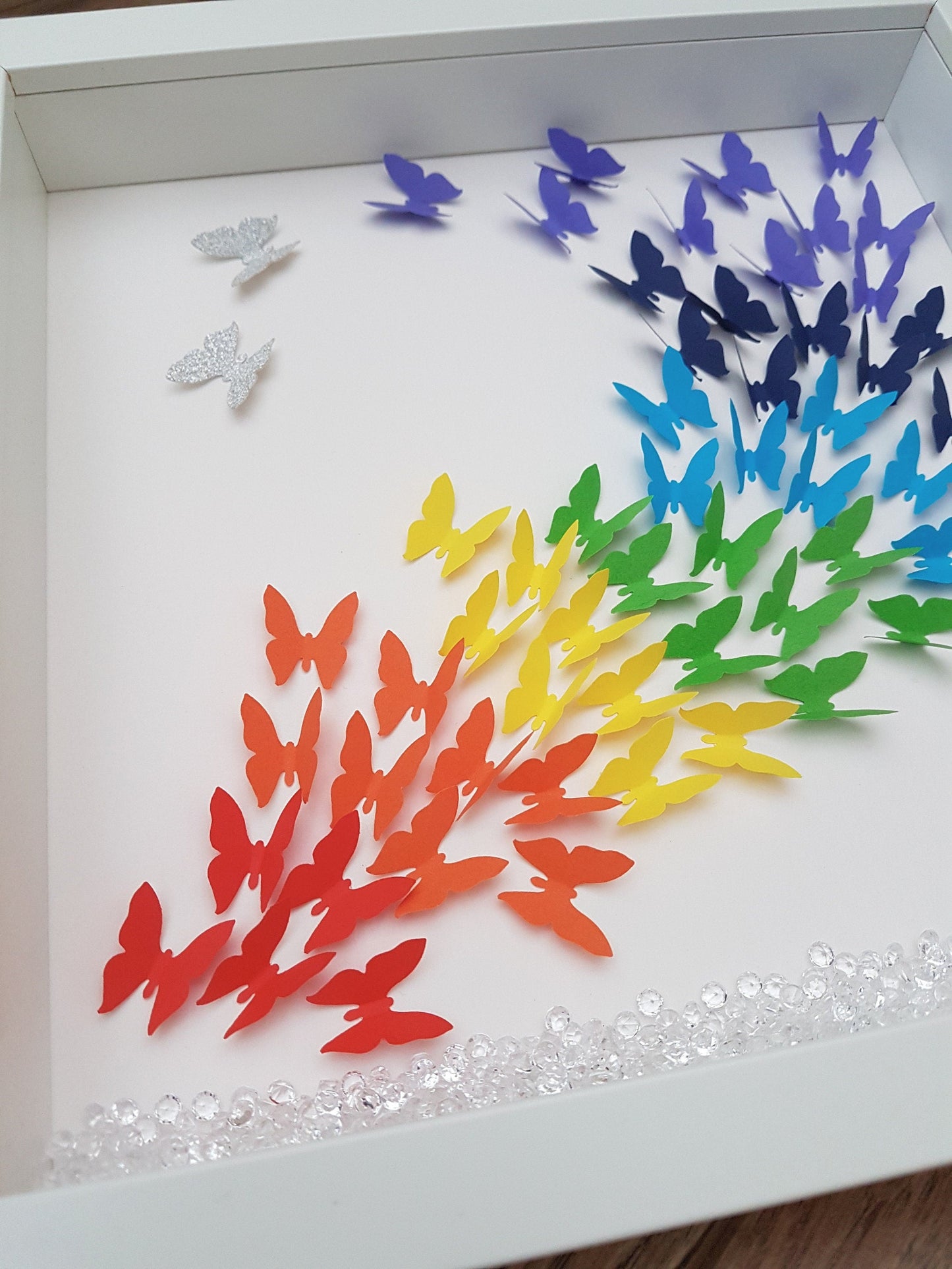 Square white box frame showing a flurry of paper butterflies in rainbow colours, moving from red butterflies in the bottom left of the frame, through rainbow colours to the top right of the frame. Plastic scatter crystals line the box frame base