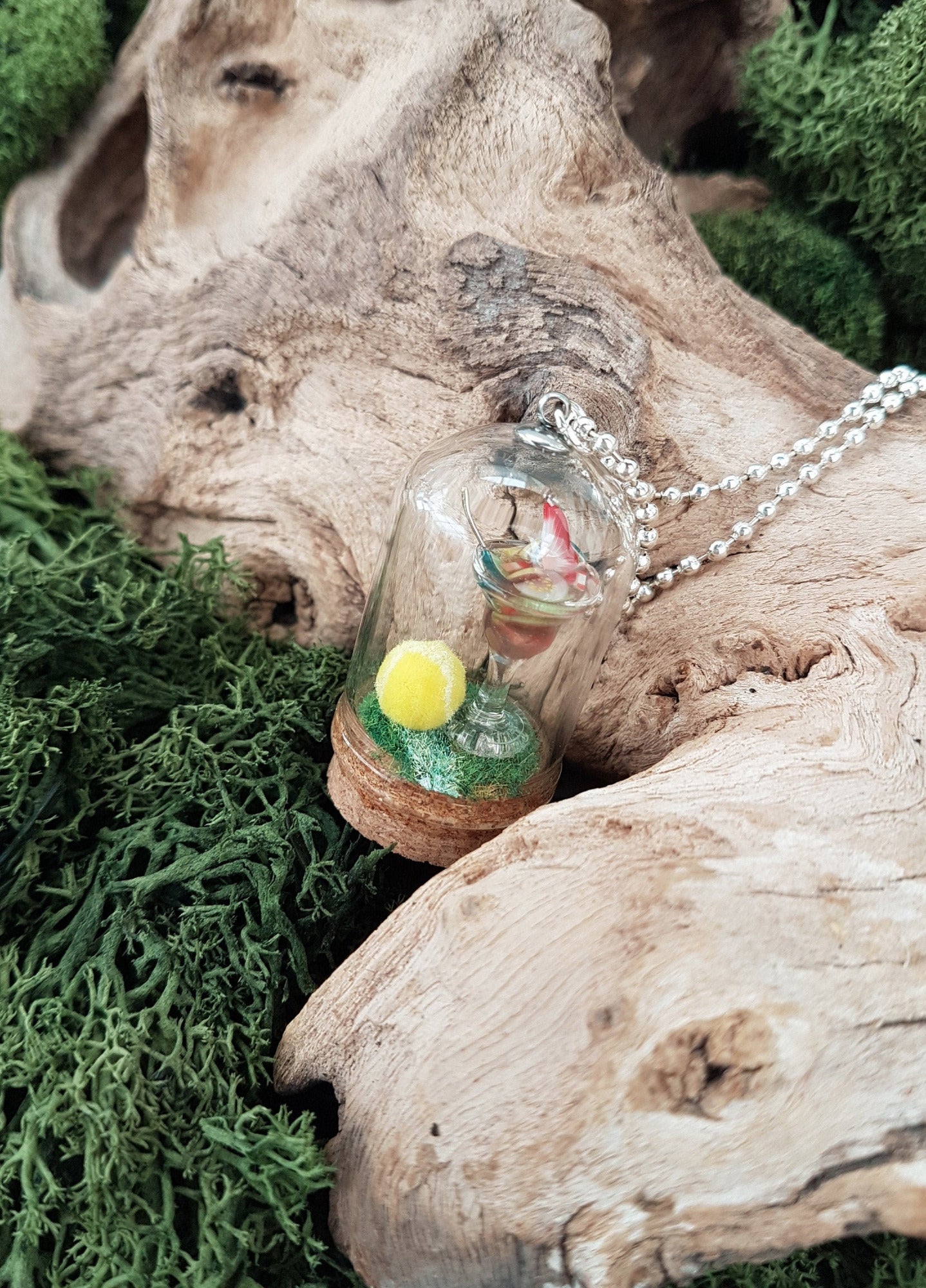 Miniature glass bell jar containing a miniature cocktail glass with Pimms and miniature fruit slices sat on a green flocked base with a fluorescent yellow tennis ball. A silver coloured chain is attached to the top of the bell jar