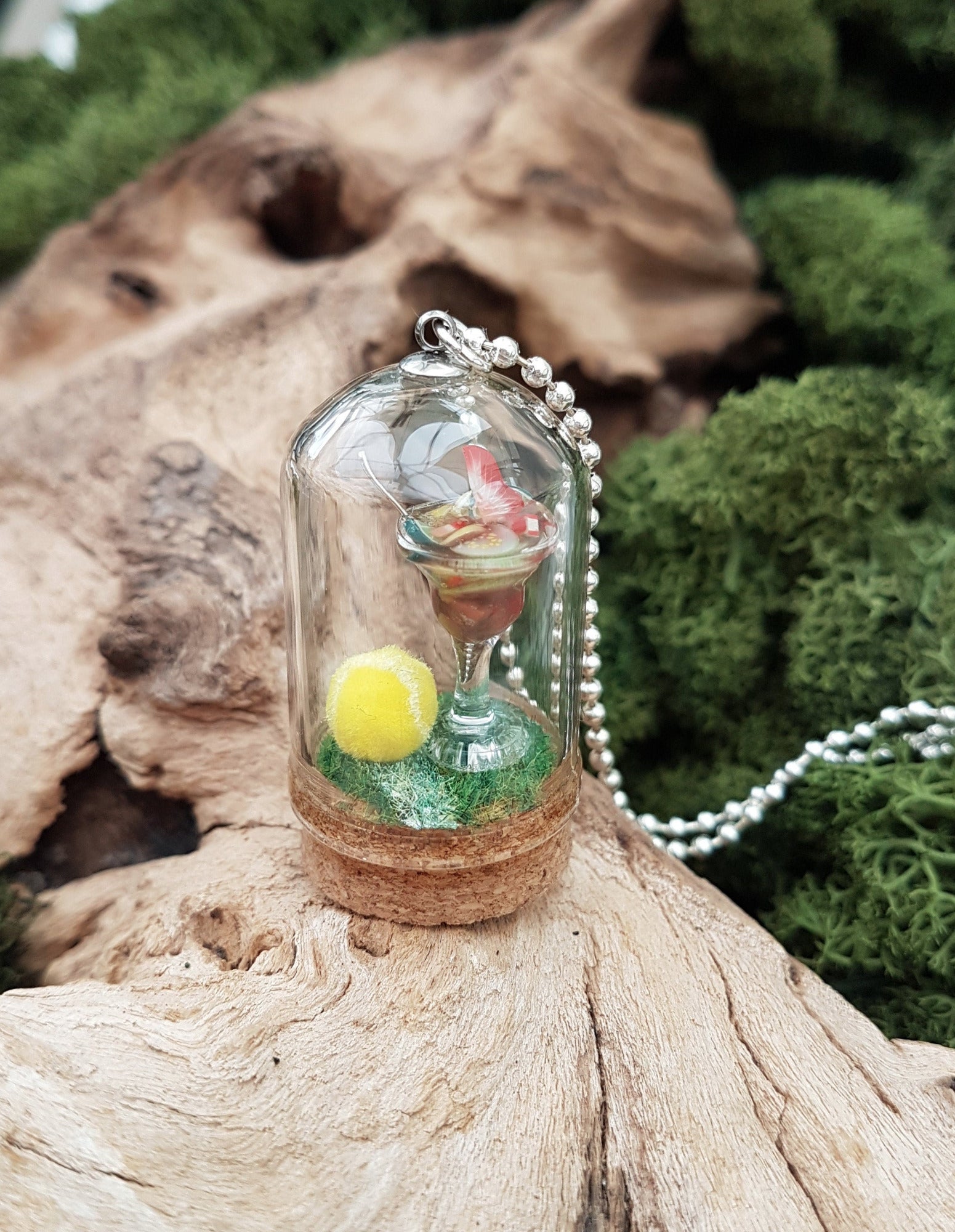 Miniature glass bell jar containing a miniature cocktail glass with Pimms and miniature fruit slices sat on a green flocked base with a fluorescent yellow tennis ball. A silver coloured chain is attached to the top of the bell jar