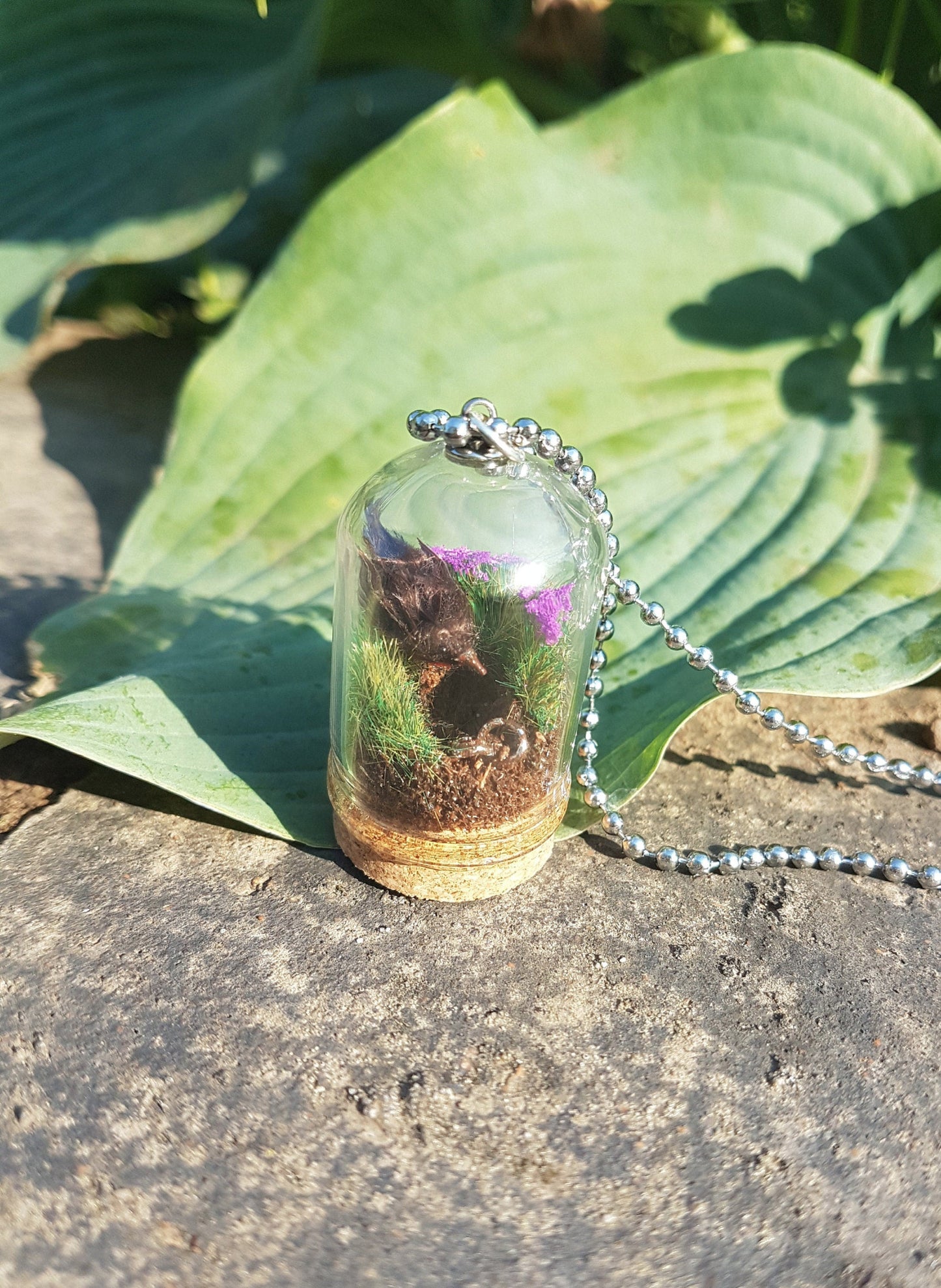 Miniature diorama glass bell jar necklace with cork base contains a tiny brown Robin. The Robin is surrounded by grass and purple flowers looking into an earthy hole containing a rusty key. The necklace is displayed on a grey stone with green leaves