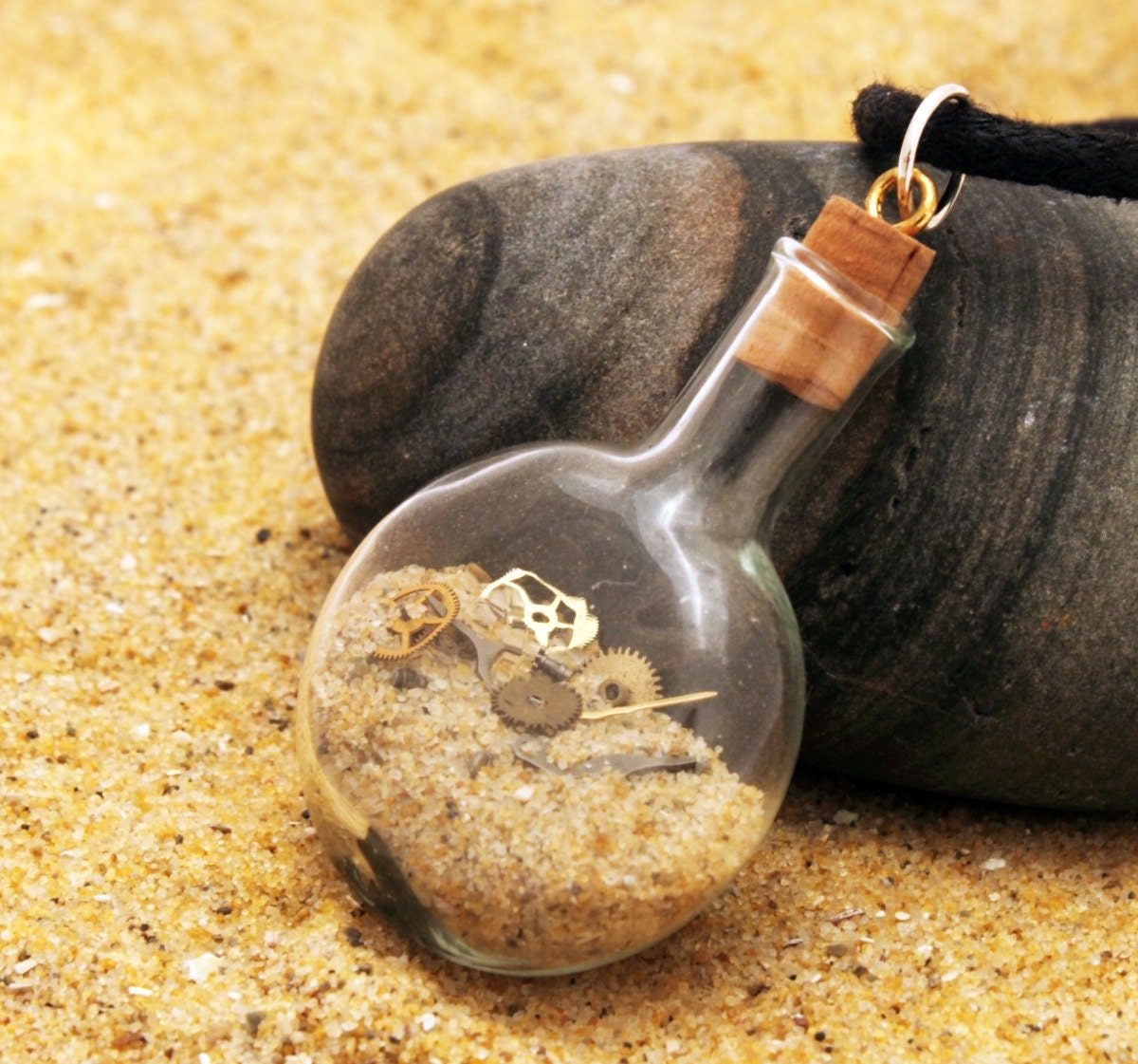 A small cognac shaped glass vial contains golden sand and tiny watch parts. Small cogs and screws can be seen within the sand. The vial is topped with a cord lid with a black cord attached. Sat on top of sand against a pebble