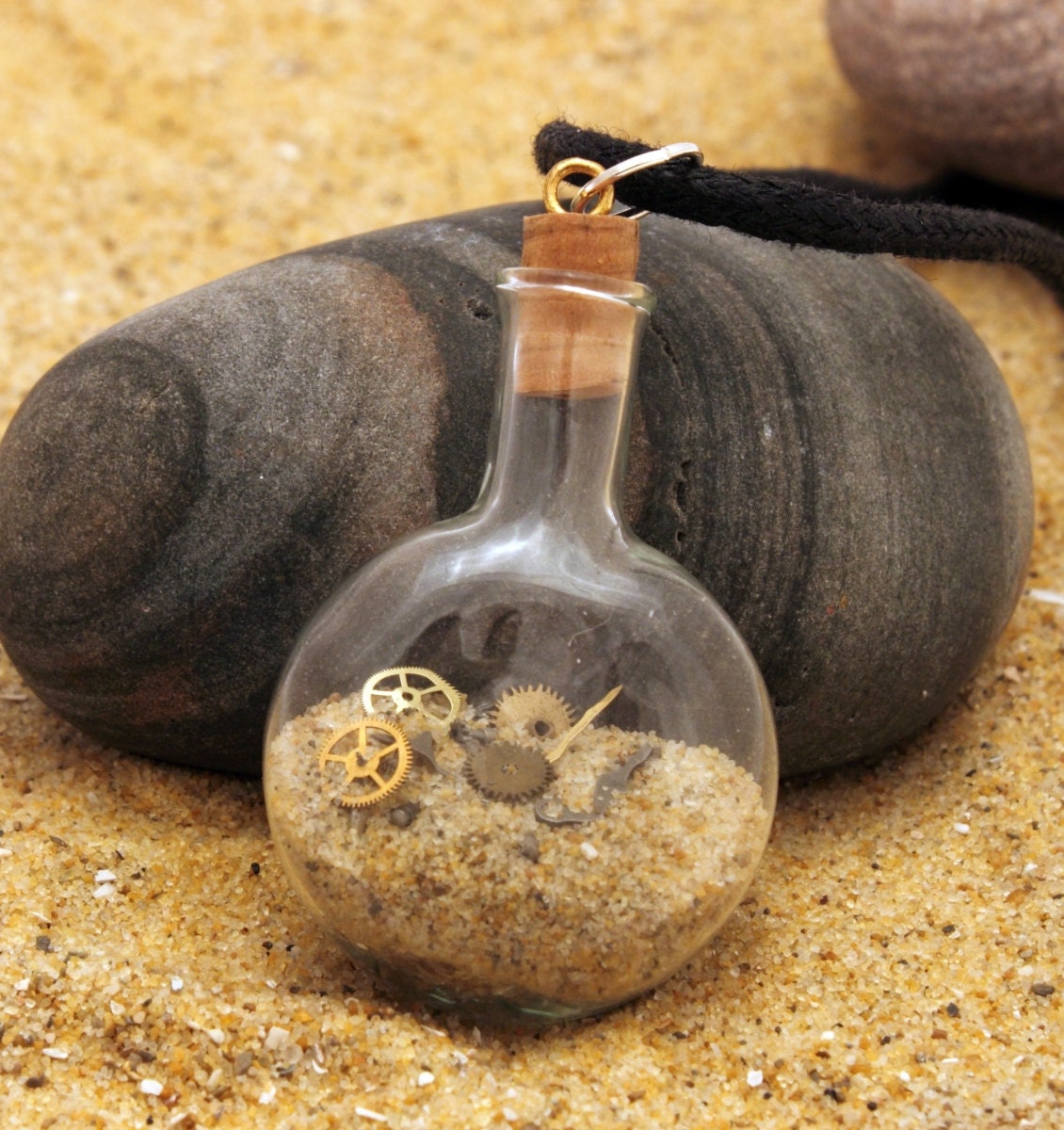 A small cognac shaped glass vial contains golden sand and tiny watch parts. Small cogs and screws can be seen within the sand. The vial is topped with a cord lid with a black cord attached. Sat on top of sand against a pebble