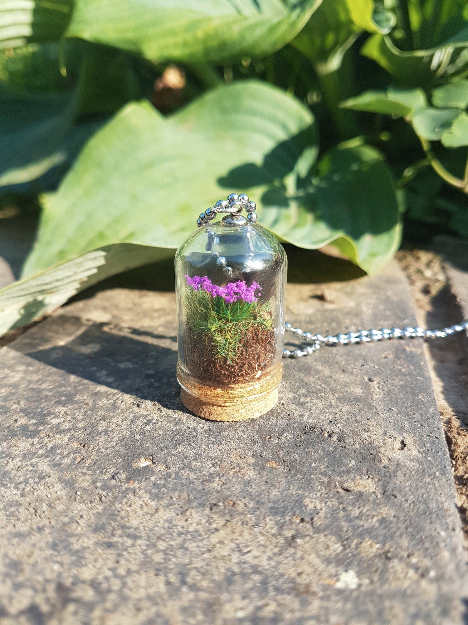 Side view of Miniature diorama glass bell jar necklace with cork base. This side shows grass and purple flowers. The necklace is displayed on a grey stone with green