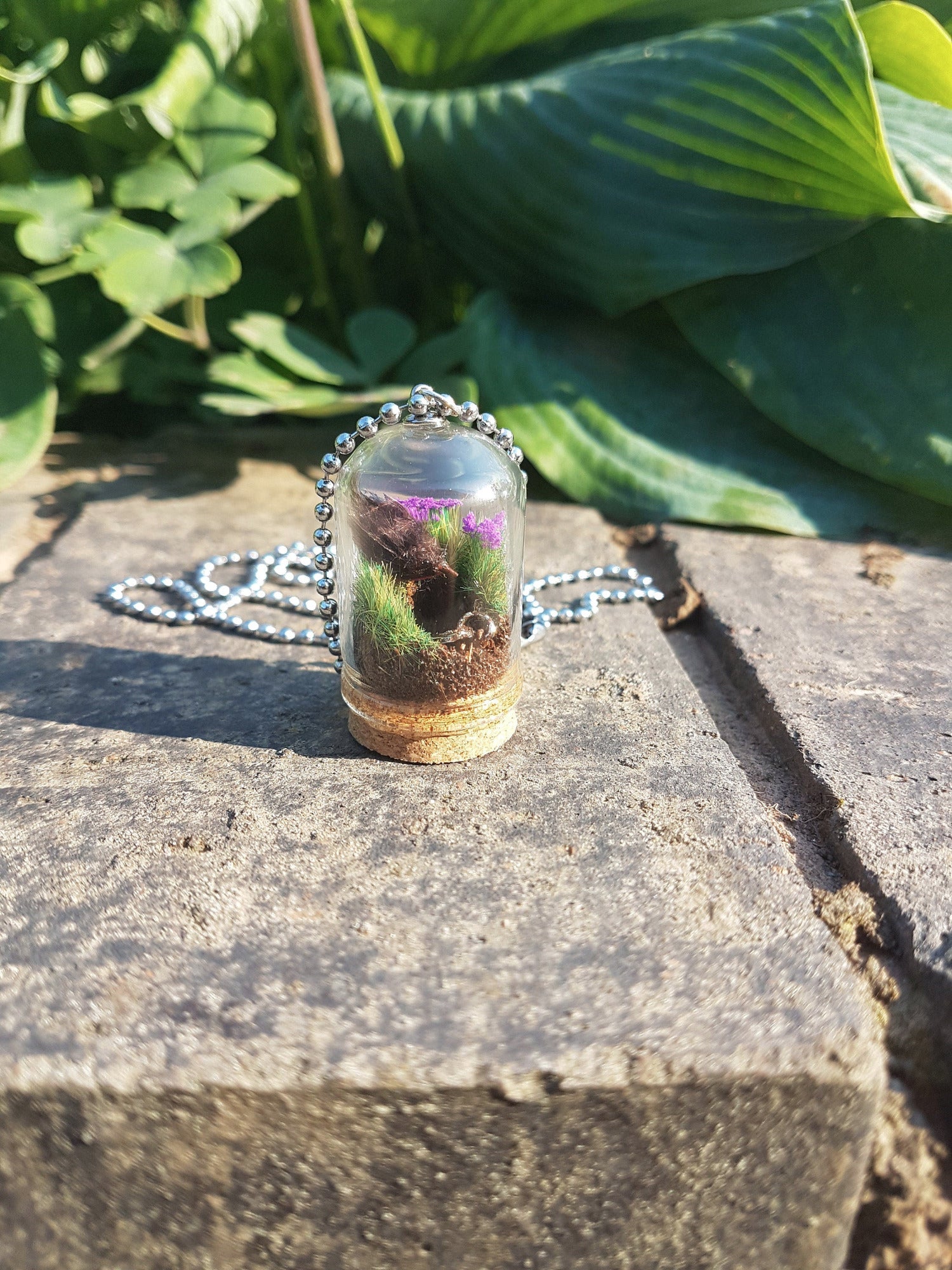 Miniature diorama glass bell jar necklace with cork base contains a tiny brown Robin. The Robin is surrounded by grass and purple flowers looking into an earthy hole containing a rusty key. The necklace is displayed on a grey stone with green leaves