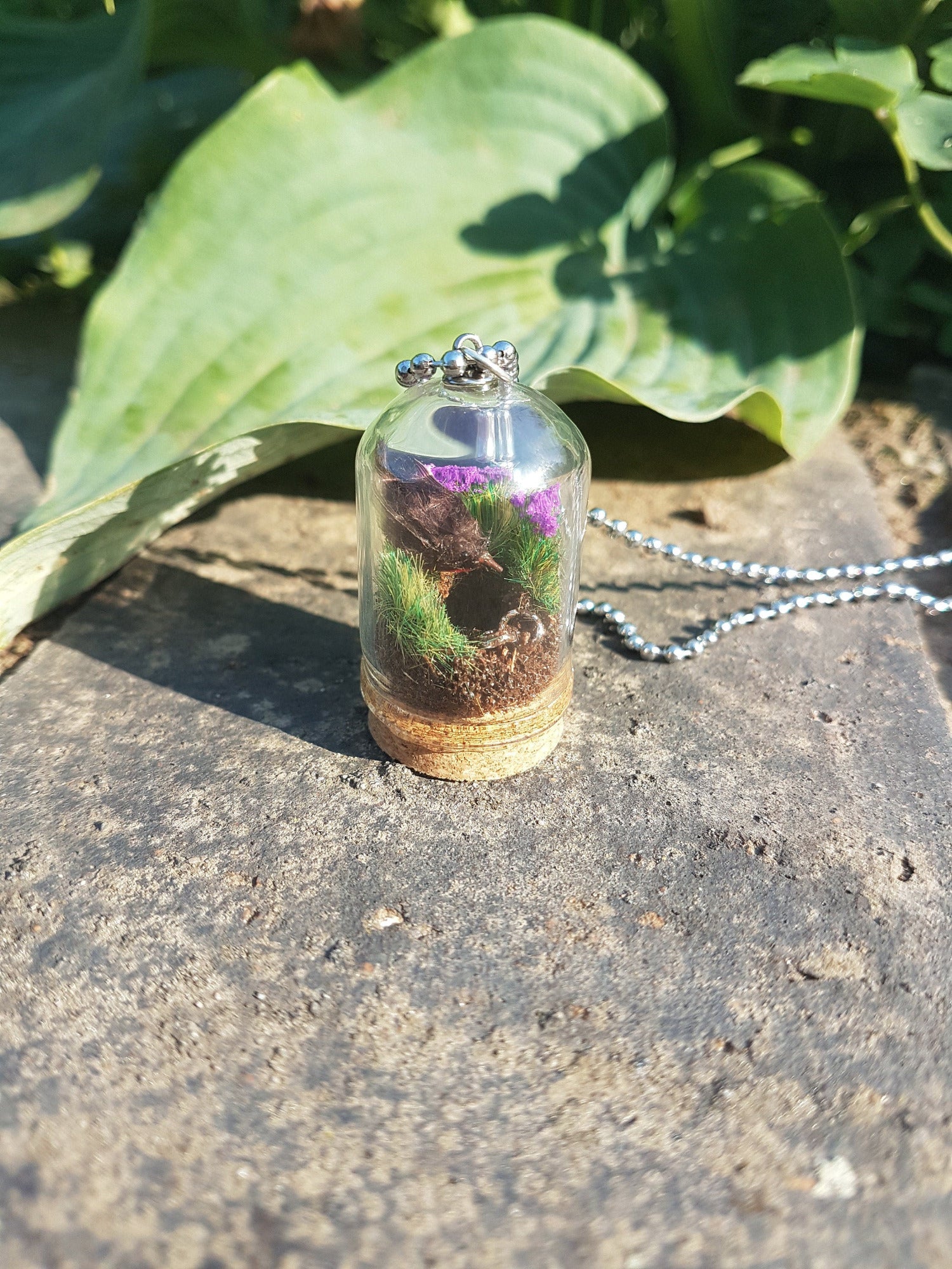 Miniature diorama glass bell jar necklace with cork base contains a tiny brown Robin. The Robin is surrounded by grass and purple flowers looking into an earthy hole containing a rusty key. The necklace is displayed on a grey stone with green leavesMiniature diorama glass bell jar necklace with cork base contains a tiny brown Robin. The Robin is surrounded by grass and purple flowers looking into an earthy hole containing a rusty key. The necklace is displayed on a grey stone with green leaves