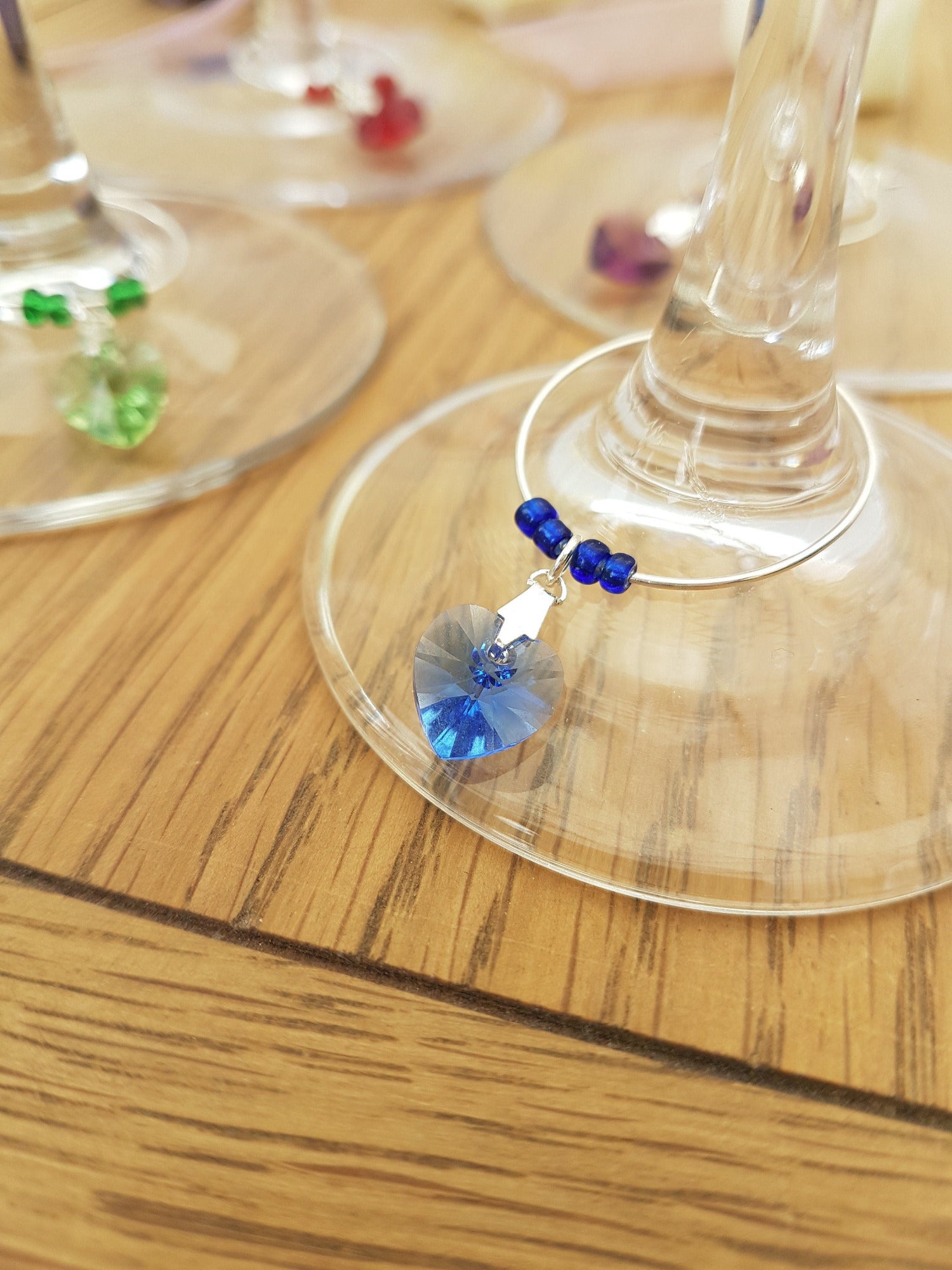 4 wine glass charms are arranged on top of a wine glass bases on a wooden background. The wine glass charms are round wire hoops, each with different coloured seed beads and a Swarovski crystal heart in red, green, blue and lavender