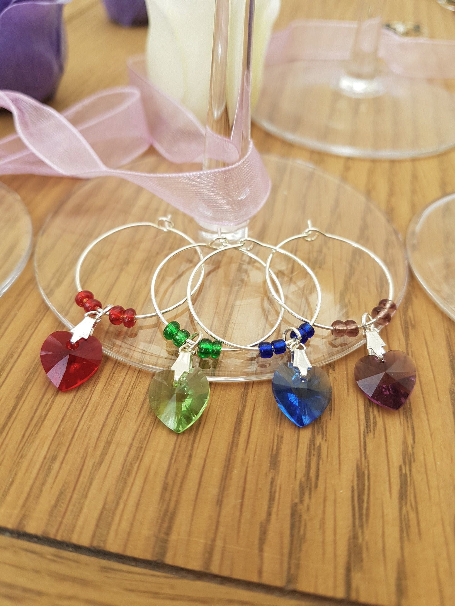 4 wine glass charms are arranged  side by side on top of a wine glass bases on a wooden background. The wine glass charms are round wire hoops, each with different coloured seed beads and a Swarovski crystal heart in red, green, blue and lavender