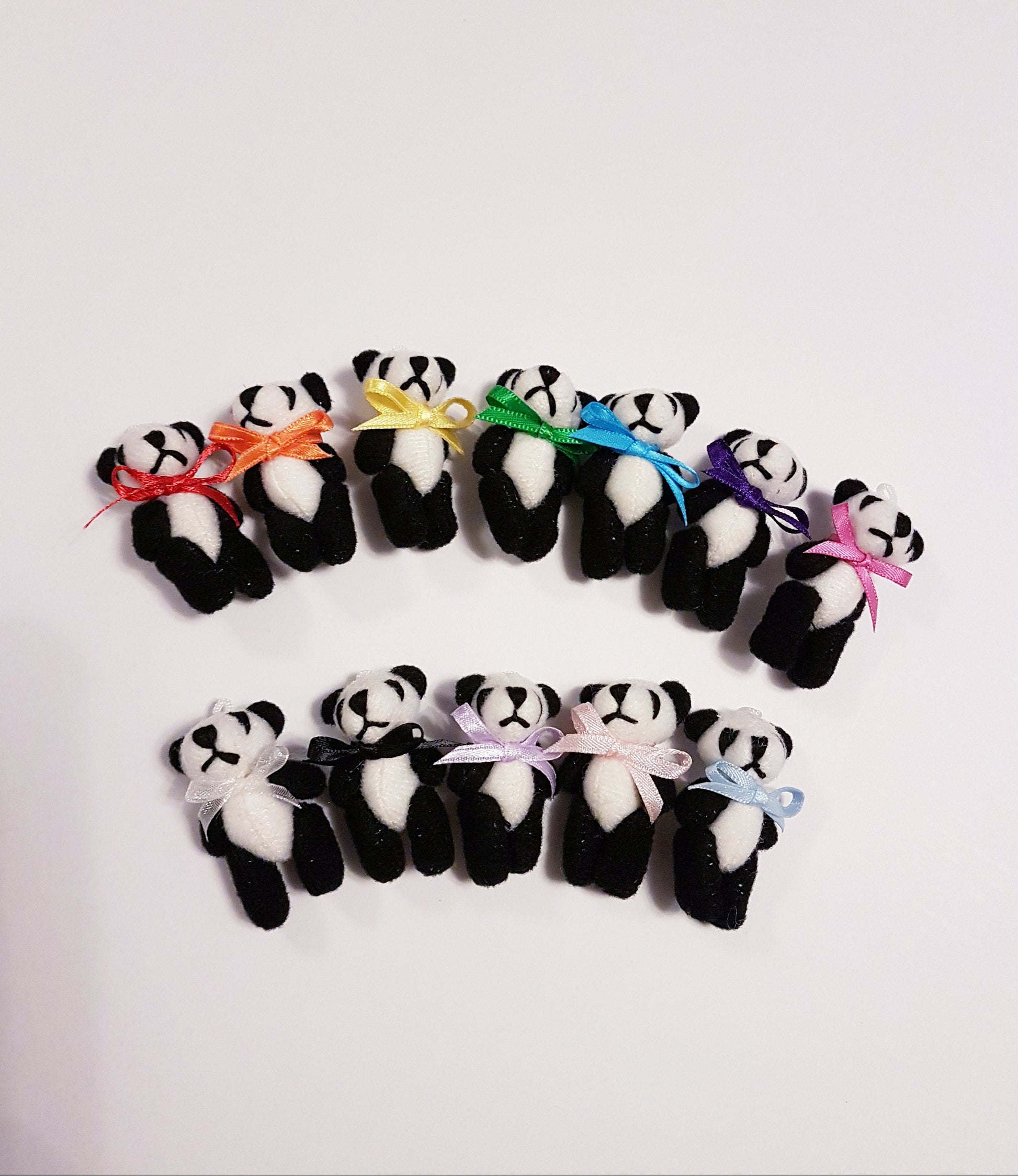 Miniature panda bears are arranged in 2 curved rows. The top row has 7 bears, each with a different coloured ribbon bowtie in rainbow colours from left to right. The bottom row has pandas with cream organza, black, pastel lilac pink and blue bow ties