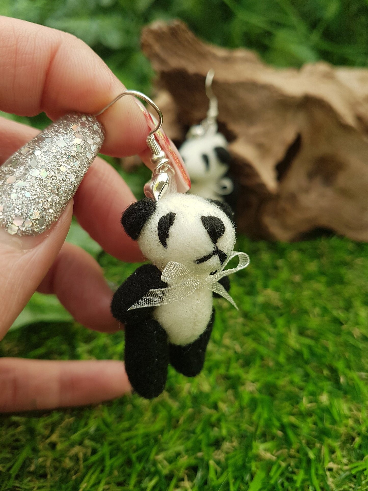 Hand holds a silver coloured hook style earring, with a small silver coloured fluffy heart charm and a larger black and cream coloured fabric panda teddy bear. The panda wears a cream organza ribbon bow tie