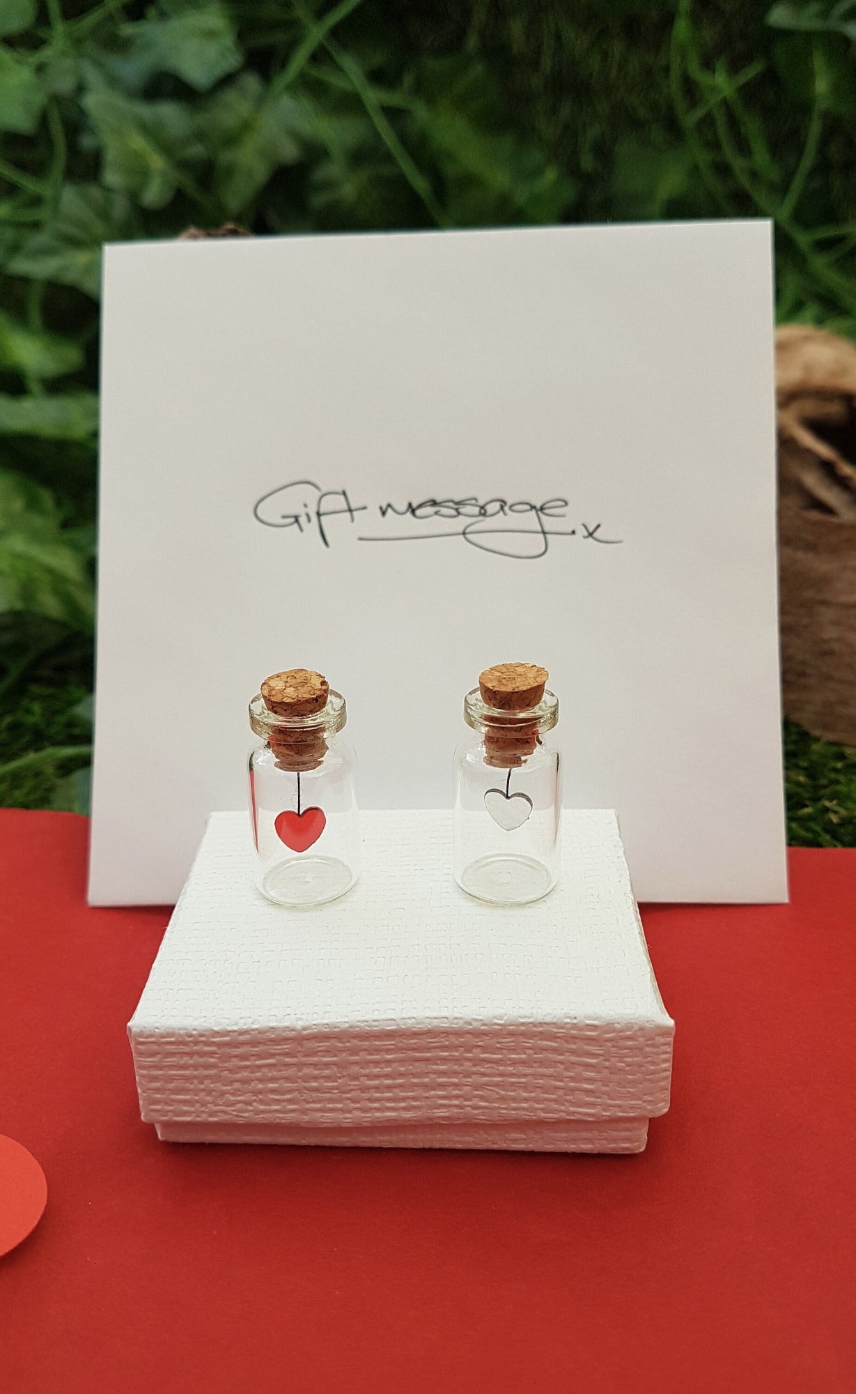 2 miniature glass jars with cork lids are sat on a white gift box on a red background. One has a red sequin heart hanging from the cork inside the jar. The other has a silver heart. A gift message envelope is in the background