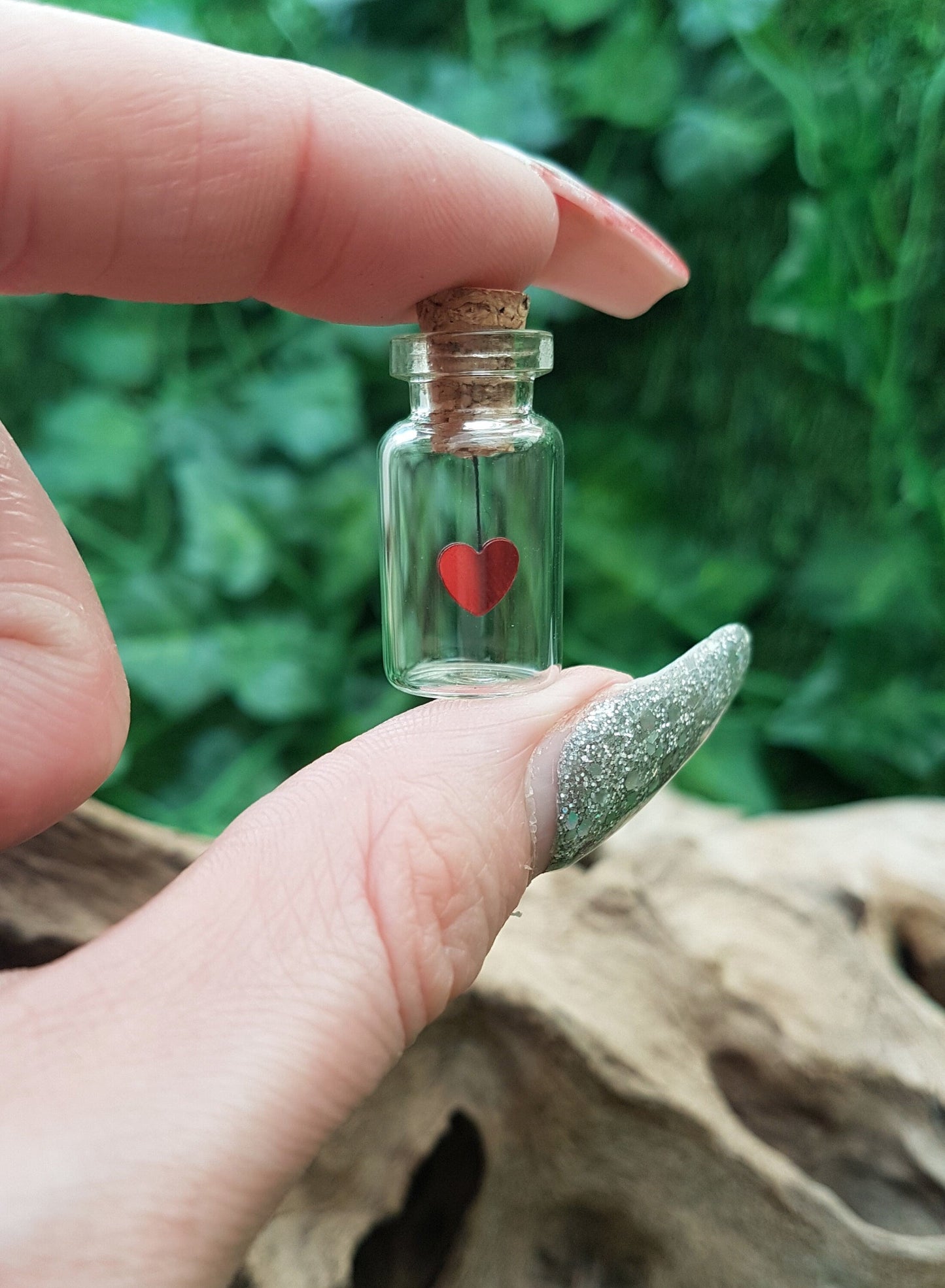 Small glass jar with cork lid with a red sequin heart hanging from the cork inside the jar, the jar is held between thumb and finger