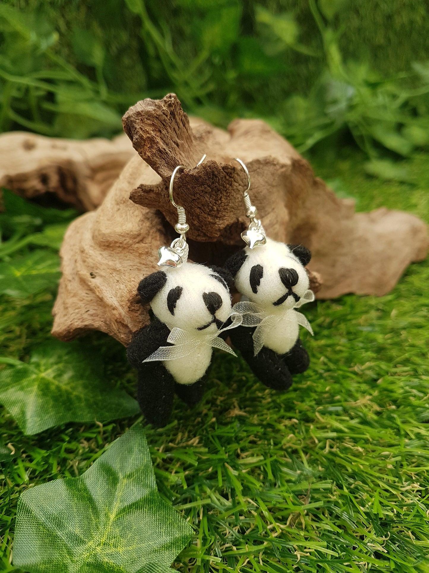 2 silver coloured hook style earrings displayed on driftwood, each with a small silver coloured fluffy heart charm and a larger black and cream coloured fabric panda teddy bear. The pandas wear a cream organza ribbon bow tie.