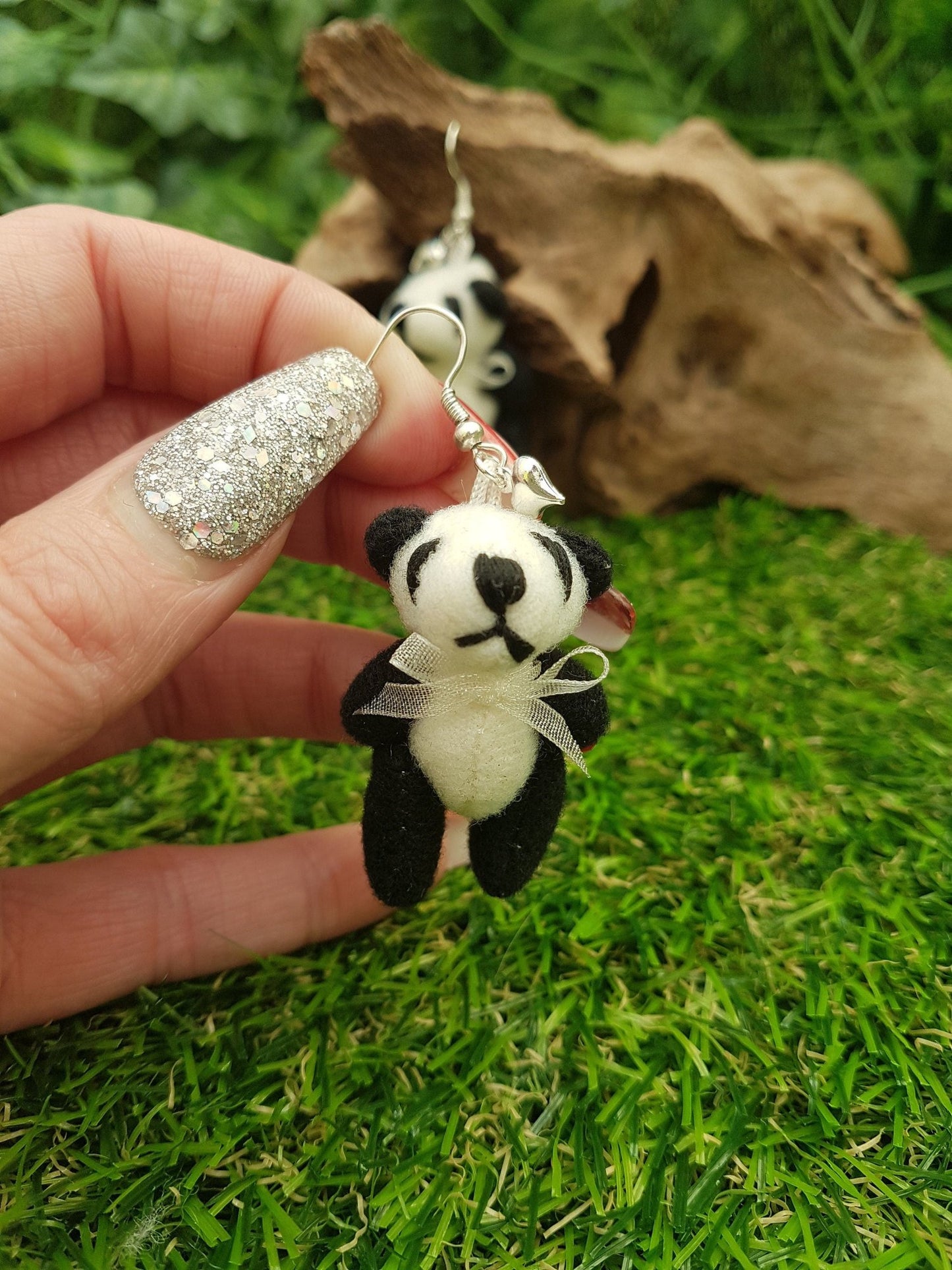 Hand holds a silver coloured hook style earrings, with a small silver coloured fluffy heart charm and a larger black and cream coloured fabric panda teddy bear. The panda wears a cream organza ribbon bow tie