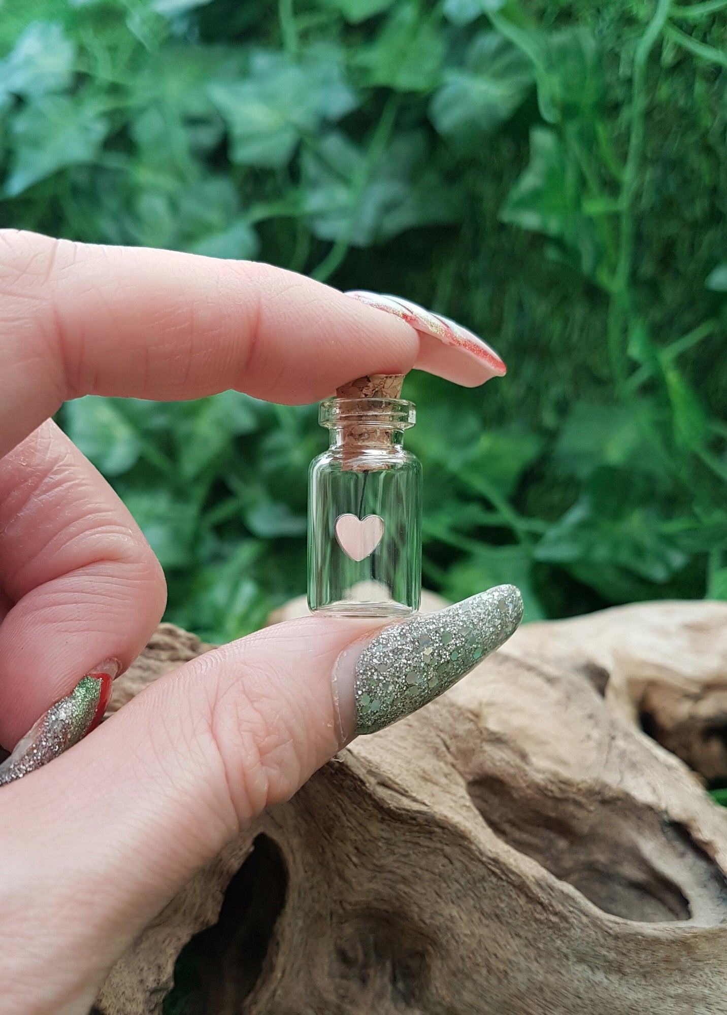 A miniature glass jar with a cork lid is held between fingers and thumb shown against a green ivy background. The jar contains a silver coloured sequin heart hanging on black cotton from the cork inside the jar