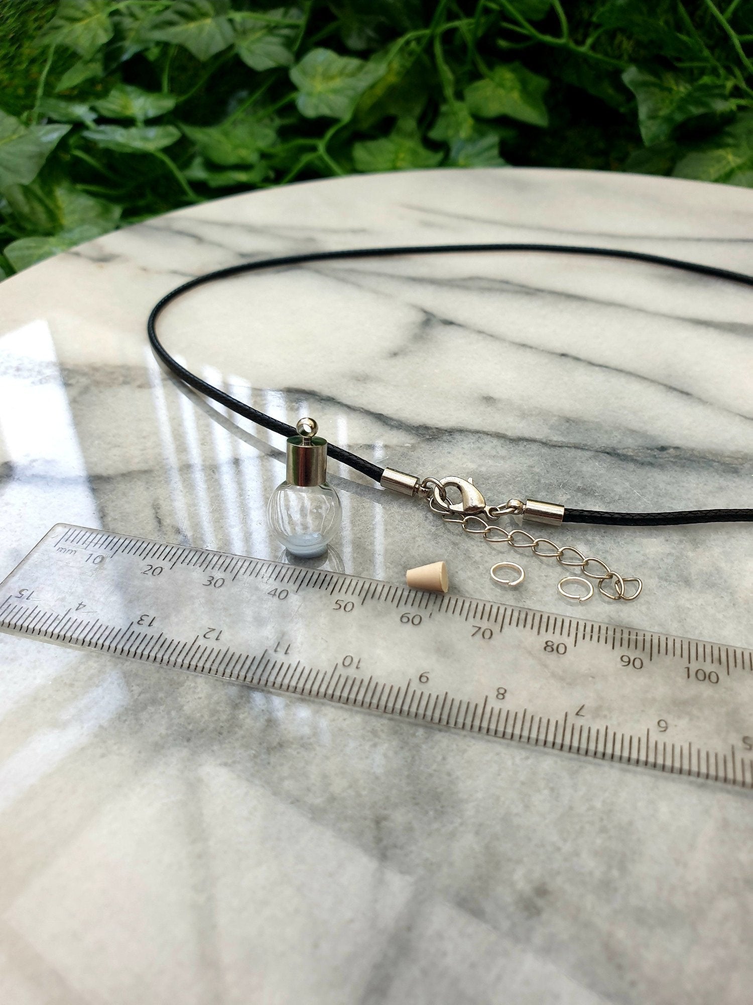 A miniature glass globe shaped vial with silver coloured cap is shown on a white marble slab with a ruler for scale. 2 jump rings, a rubber bung and a black pendant cord seen in the background.