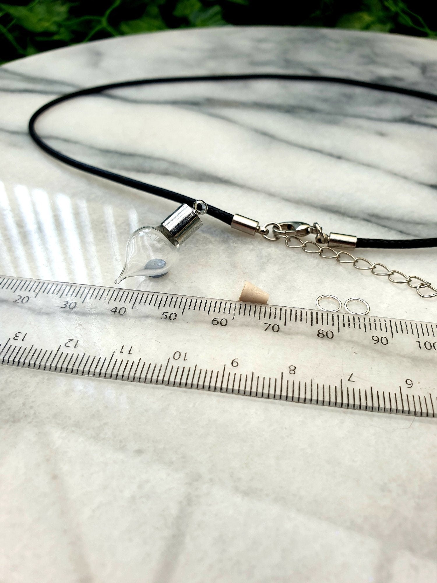 A miniature glass vial with silver coloured caps shown on a white marble slab with a ruler for scale. The vial looks like an amphora, rounded with a pointed base. 2 jump rings, a rubber bung and a black pendant cord seen in the background.