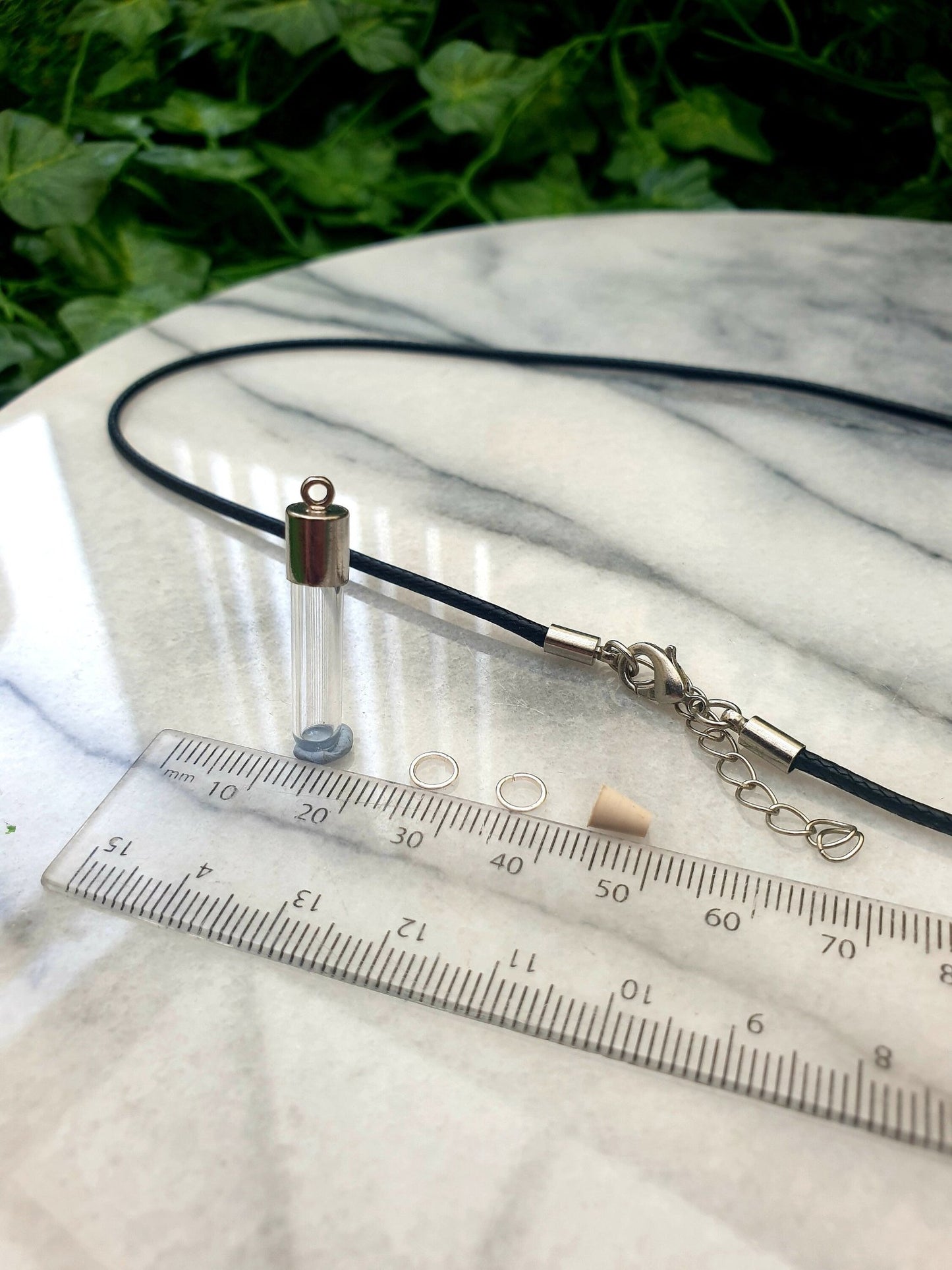 A miniature glass test tube shaped vial with silver coloured cap is shown on a white marble slab with a ruler for scale. 2 jump rings, a rubber bung and a black pendant cord seen in the background.