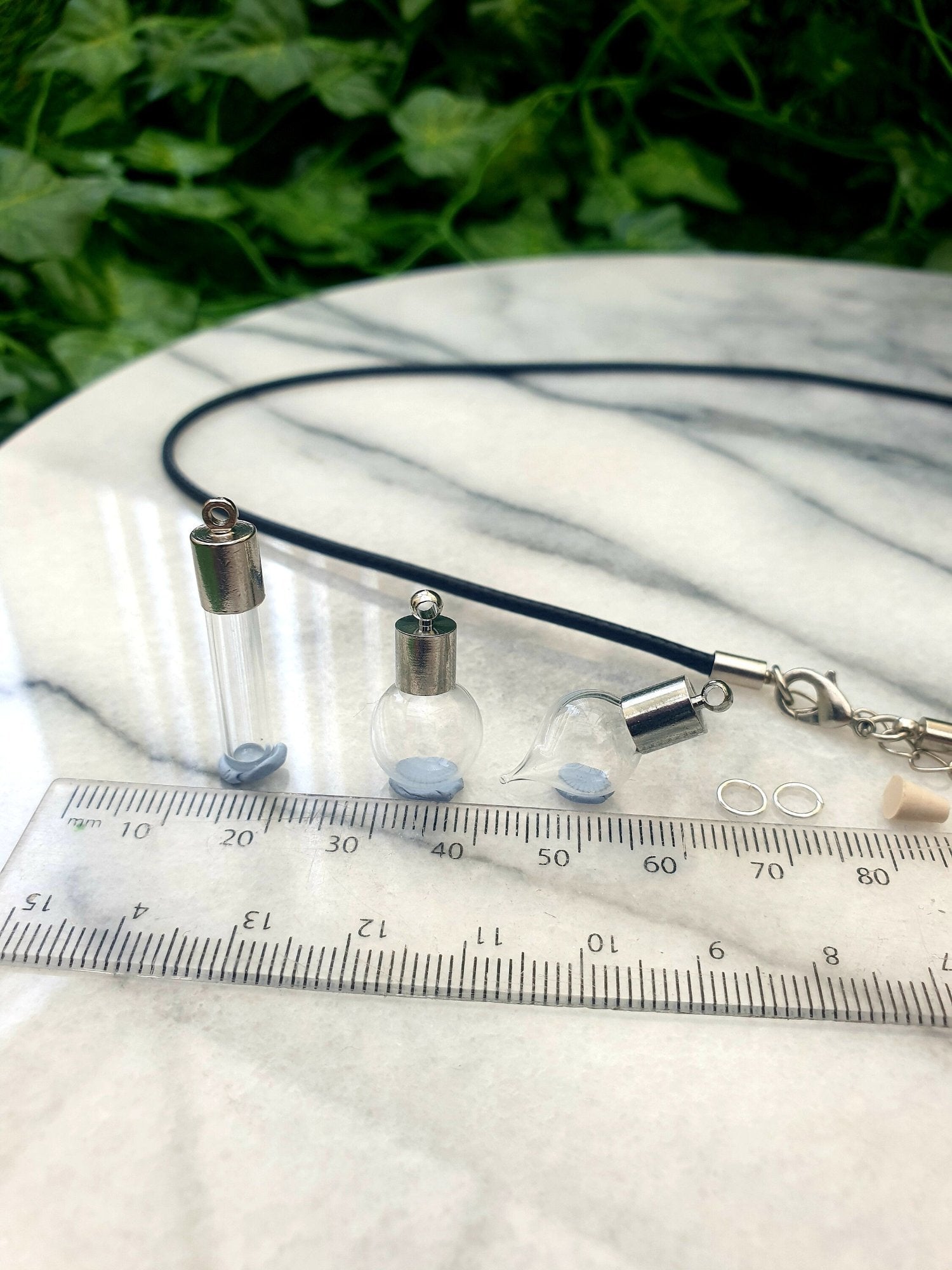 3 miniature glass vials with silver coloured caps shown on a white marble slab with a ruler for scale. 2 jump rings, a rubber bung and a black pendant cord seen in the background. One vial is tube shaped, one is rounded, one is has a pointed bottom