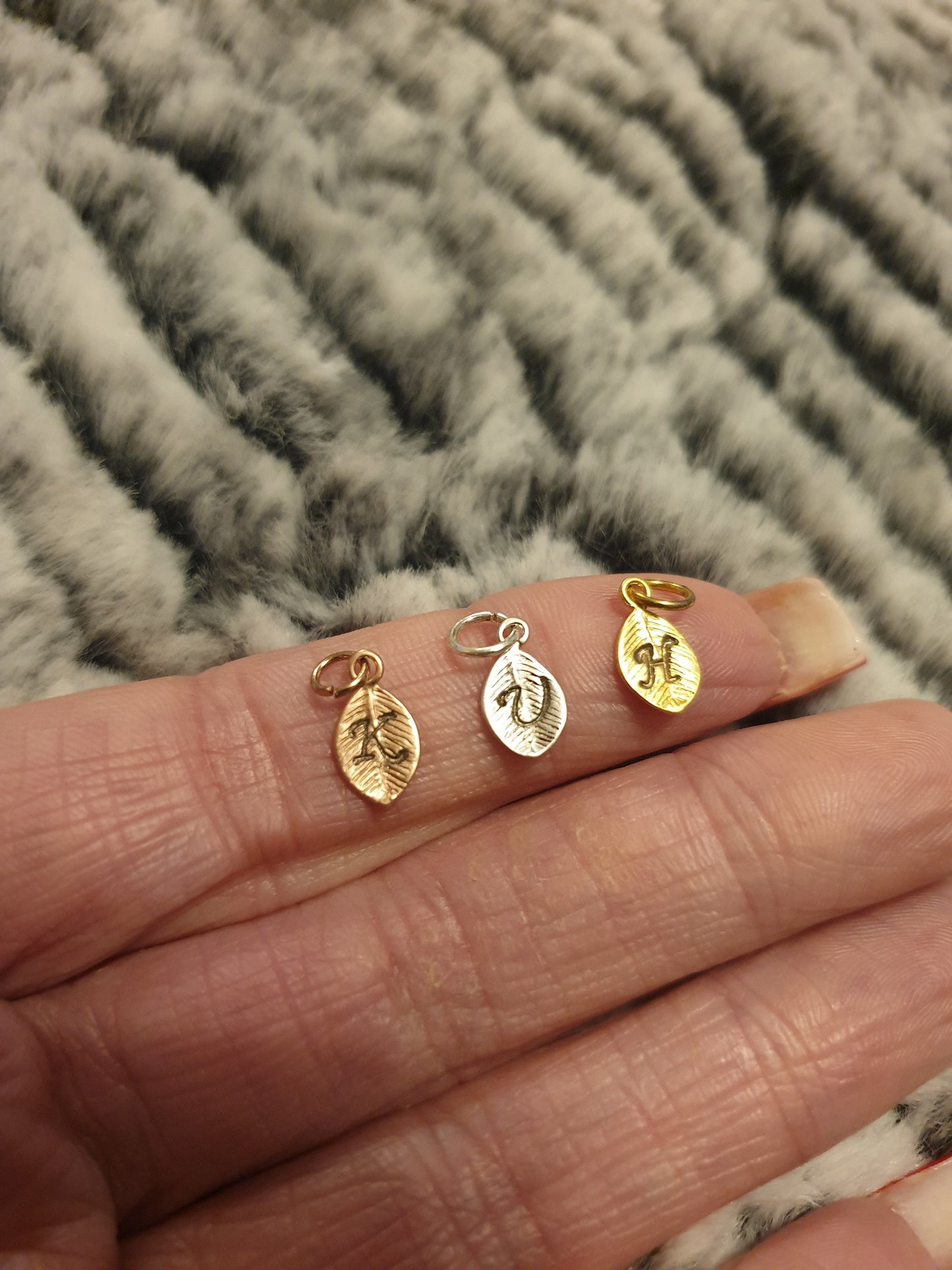 3 metal leaf-shaped charms with a small loop and jump ring. The first is rose gold coloured with a calligraphic K initial. The second is silver coloured with a V initial. The third is gold coloured with an H initial. Held on an index finger