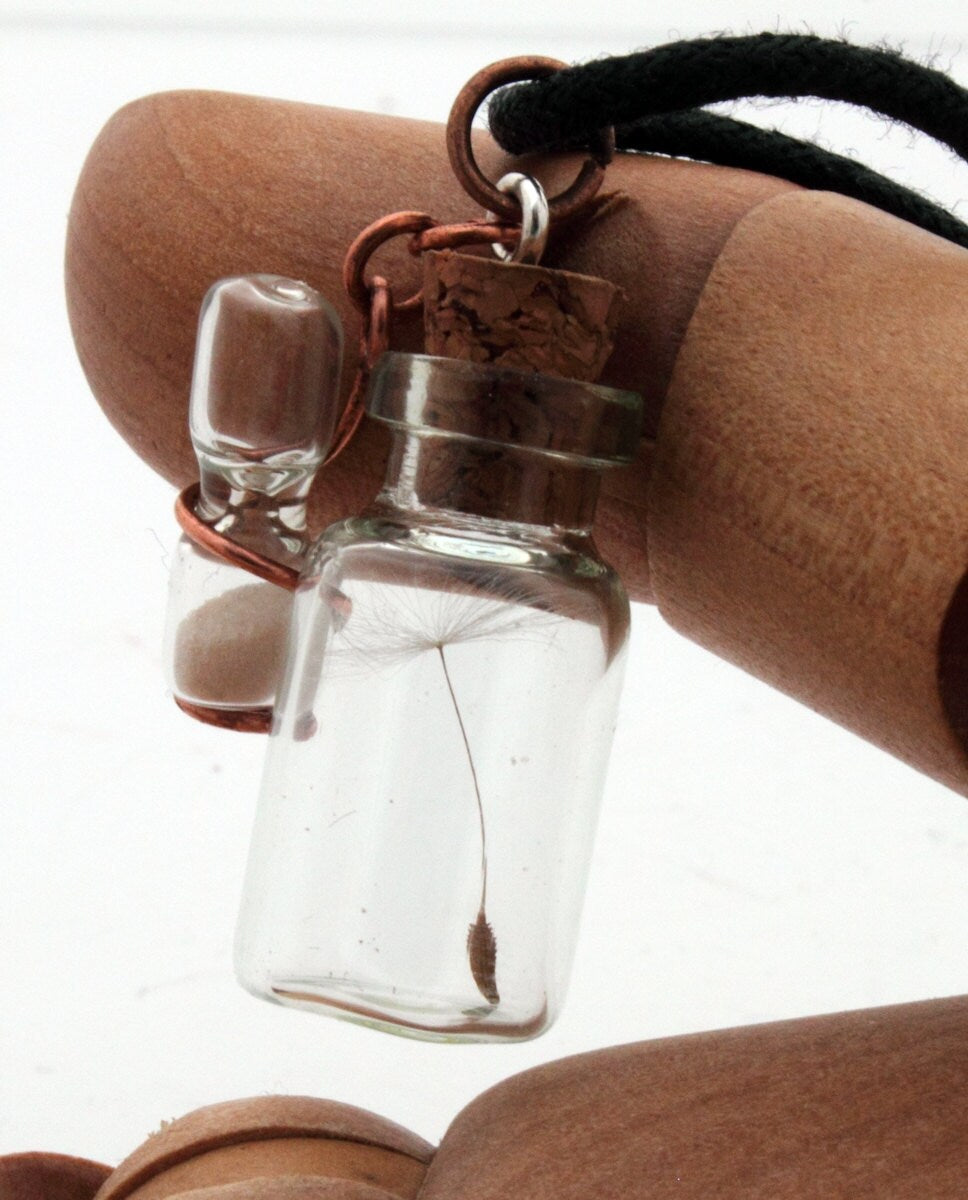 Miniature jar shaped glass vial containing a dandelion seed with a copper wrapped miniature glass sand timer attachdd to the cork lid. Displayed on a black coloured cord attached to the cork lid