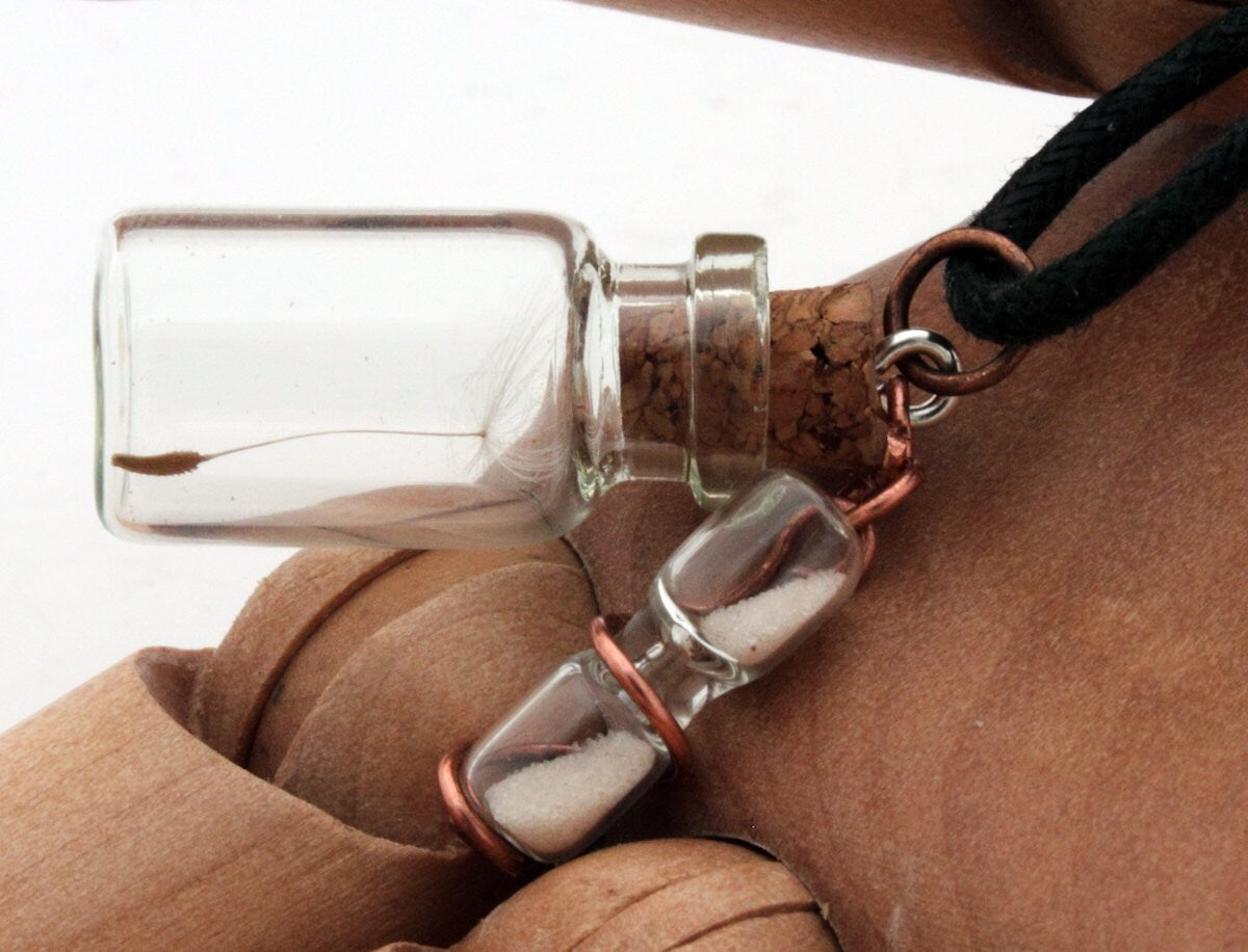 Miniature jar shaped glass vial containing a dandelion seed with a copper wrapped miniature glass sand timer attachdd to the cork lid. Displayed on a black coloured cord attached to the cork lid