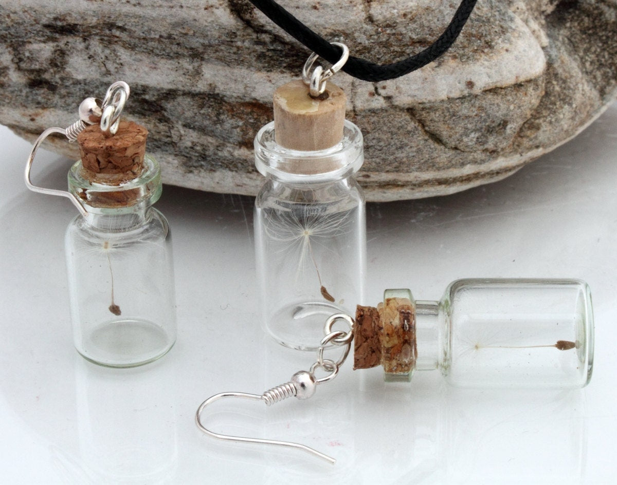 Two small glass jar vials are shown against a natural stone background. One vial is stood up and one vial is in its side, both containing dandelions clock seeds. Each jar has a cork lid with an earring hook attached. Shown with matching necklace